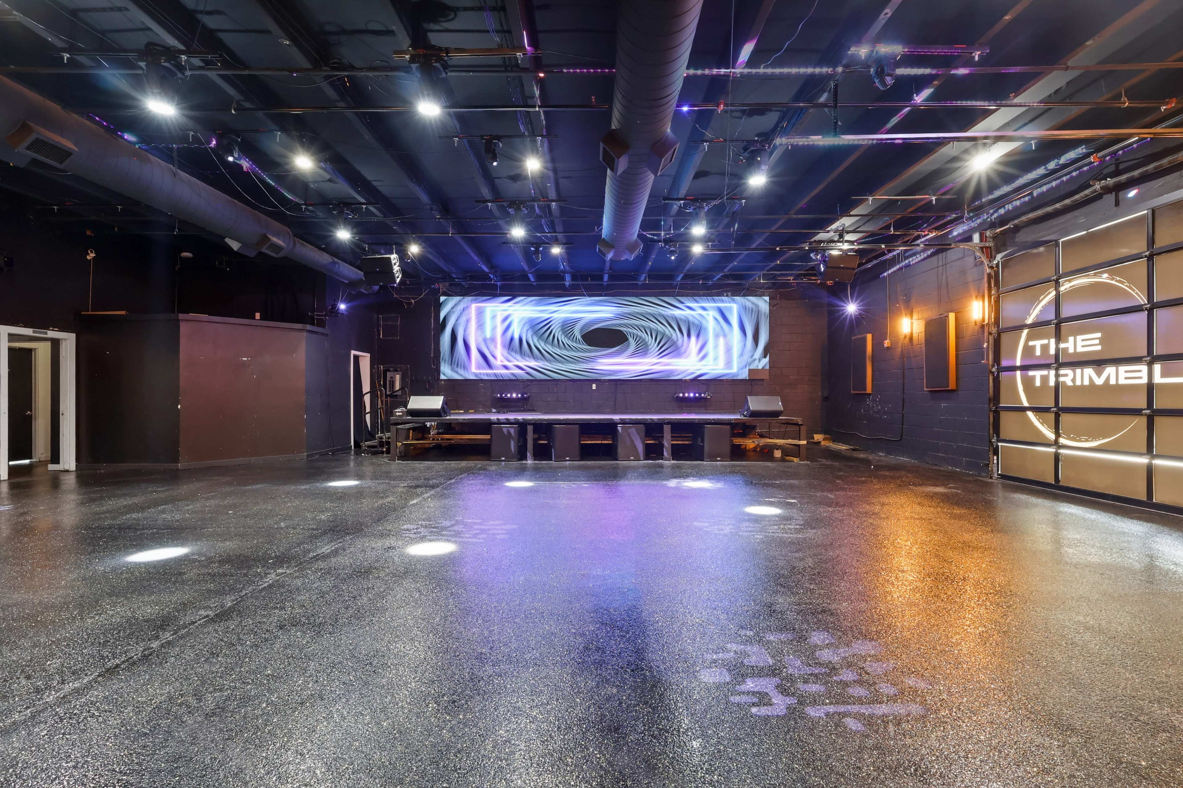An empty event space features a sleek black floor, illuminated by bright overhead lights and a large abstract display on the back wall.