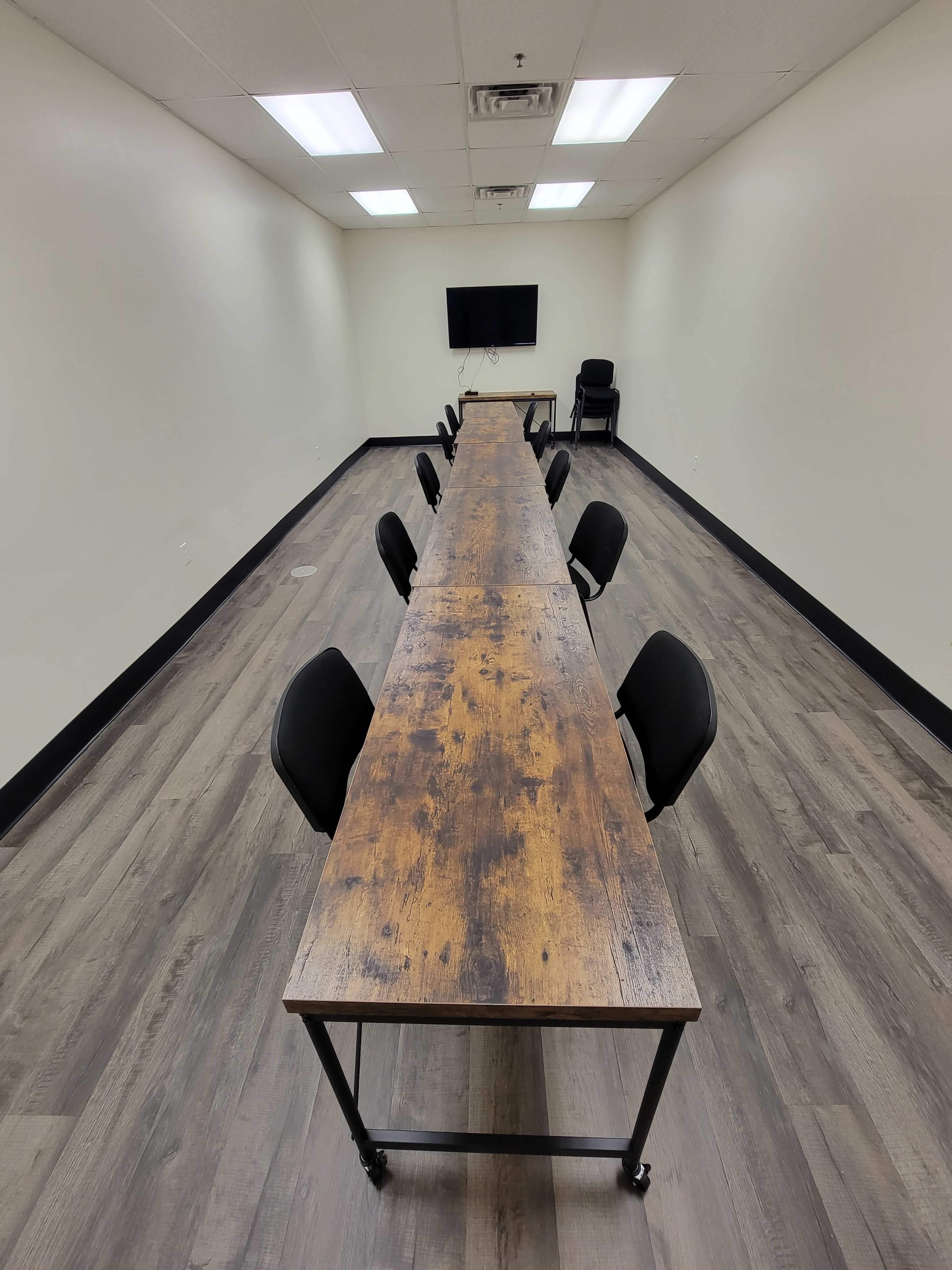 A long wooden table with black chairs is arranged in a meeting room, featuring a television mounted on the wall.
