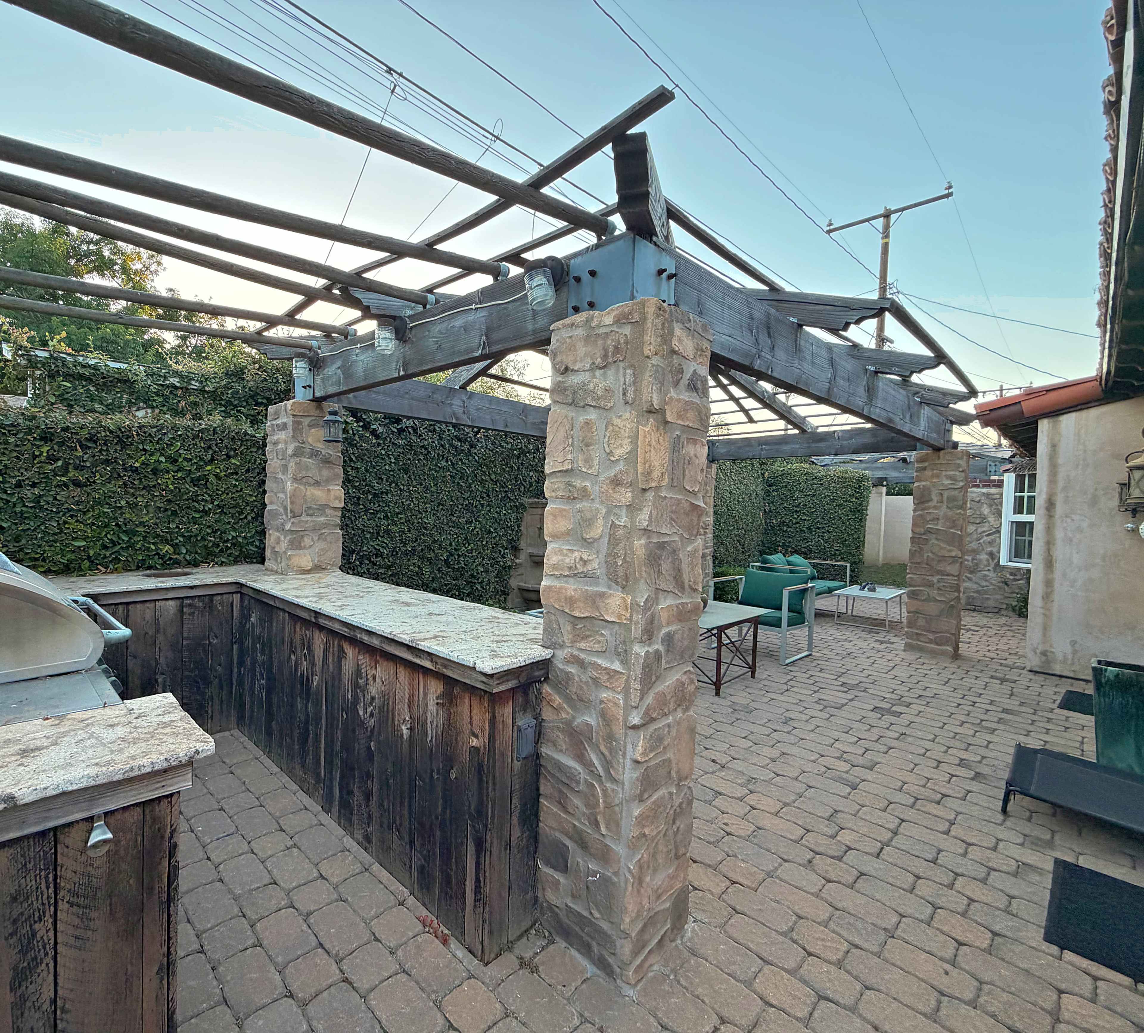The image shows a stone and wood outdoor patio area with a bar structure and seating, surrounded by greenery.