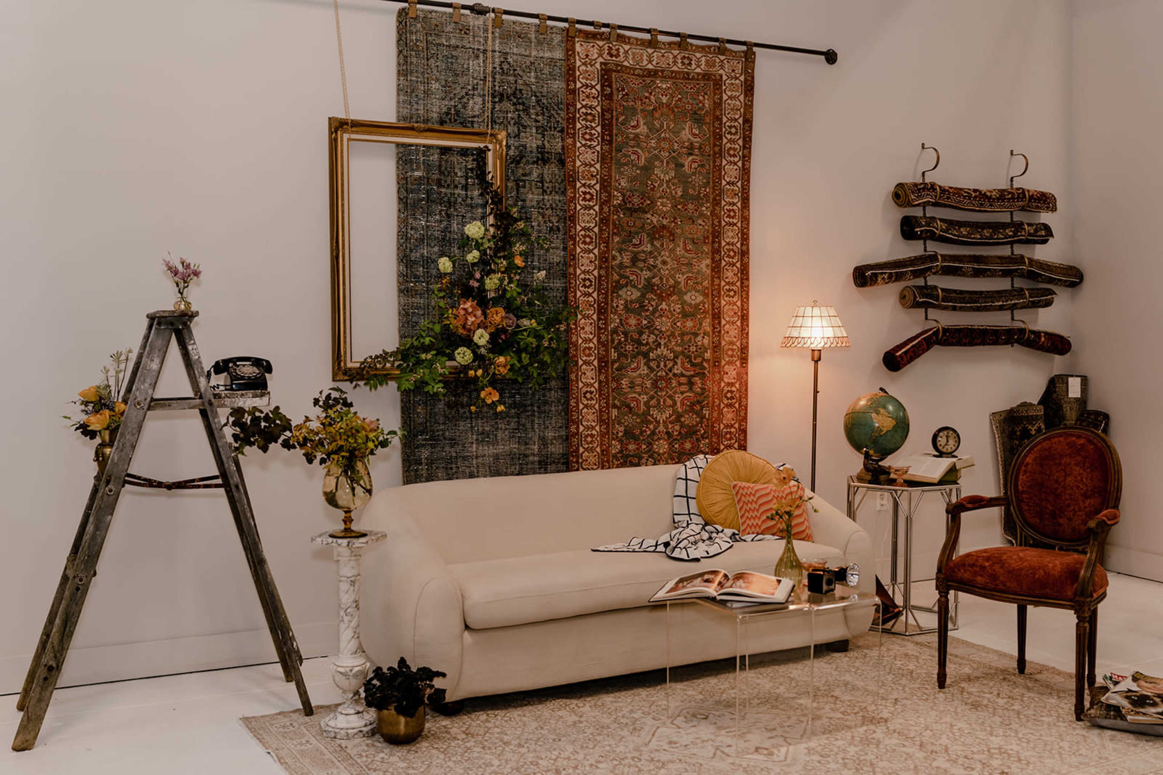The image shows a decorated living room featuring a beige sofa, a vintage armchair, a ladder with flowers, and various decorative elements on the walls.