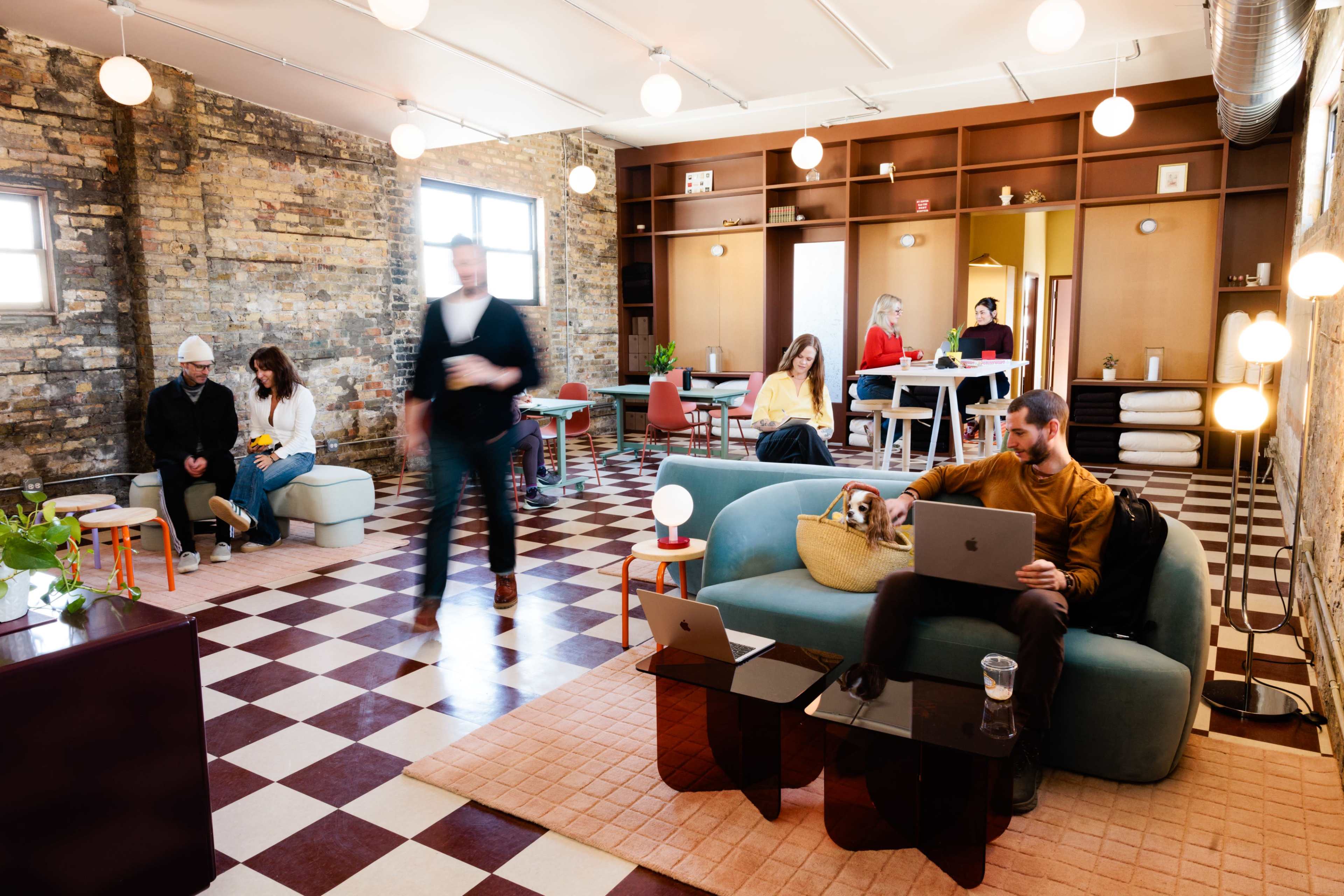 The image shows a spacious co-working area with various seating arrangements, including people working on laptops and others socializing, set against a backdrop of exposed brick walls and patterned flooring.