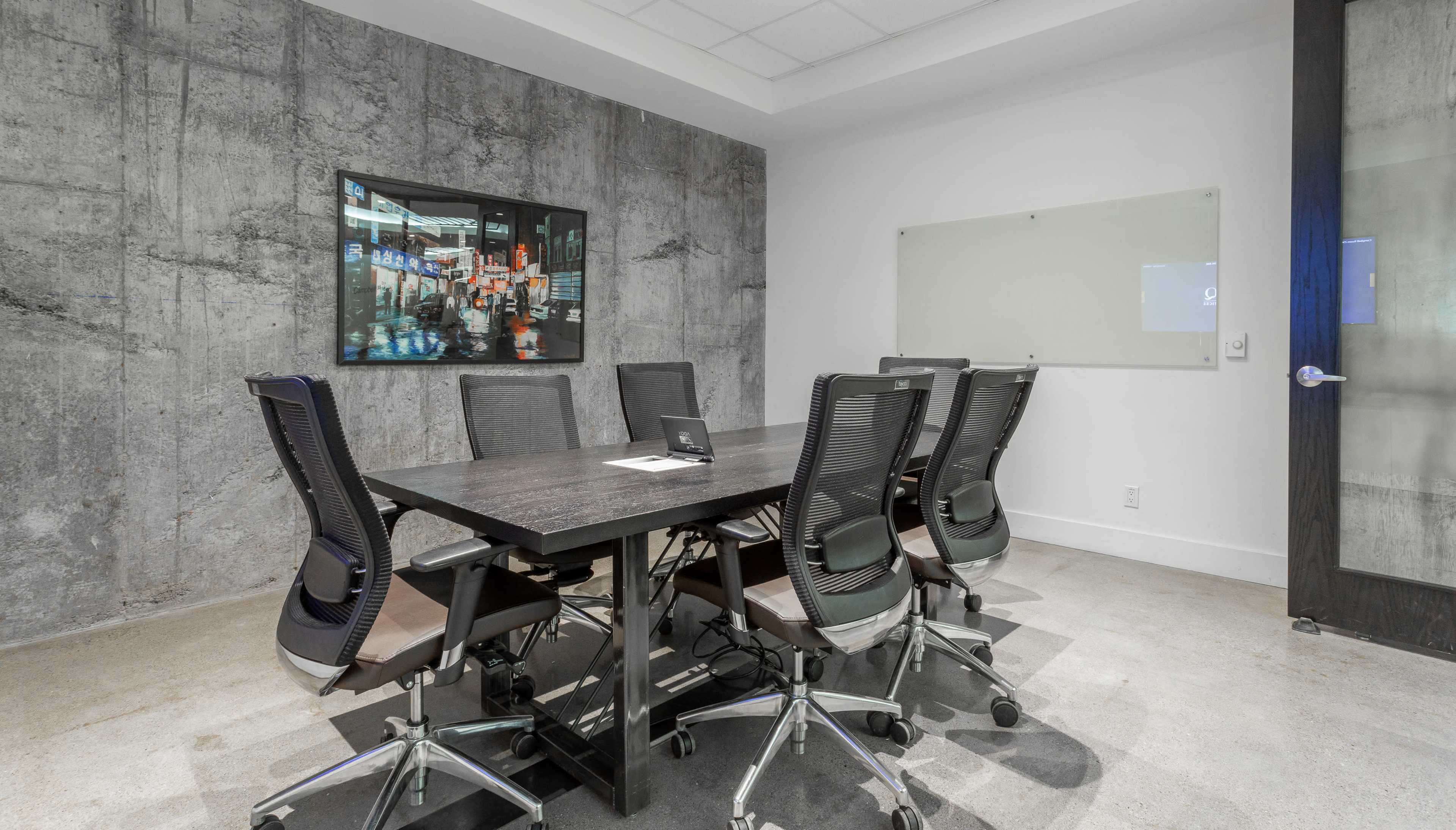 The image shows a conference room with a large table surrounded by six ergonomic chairs and a wall-mounted screen displaying a digital collage, set against a concrete-textured backdrop.