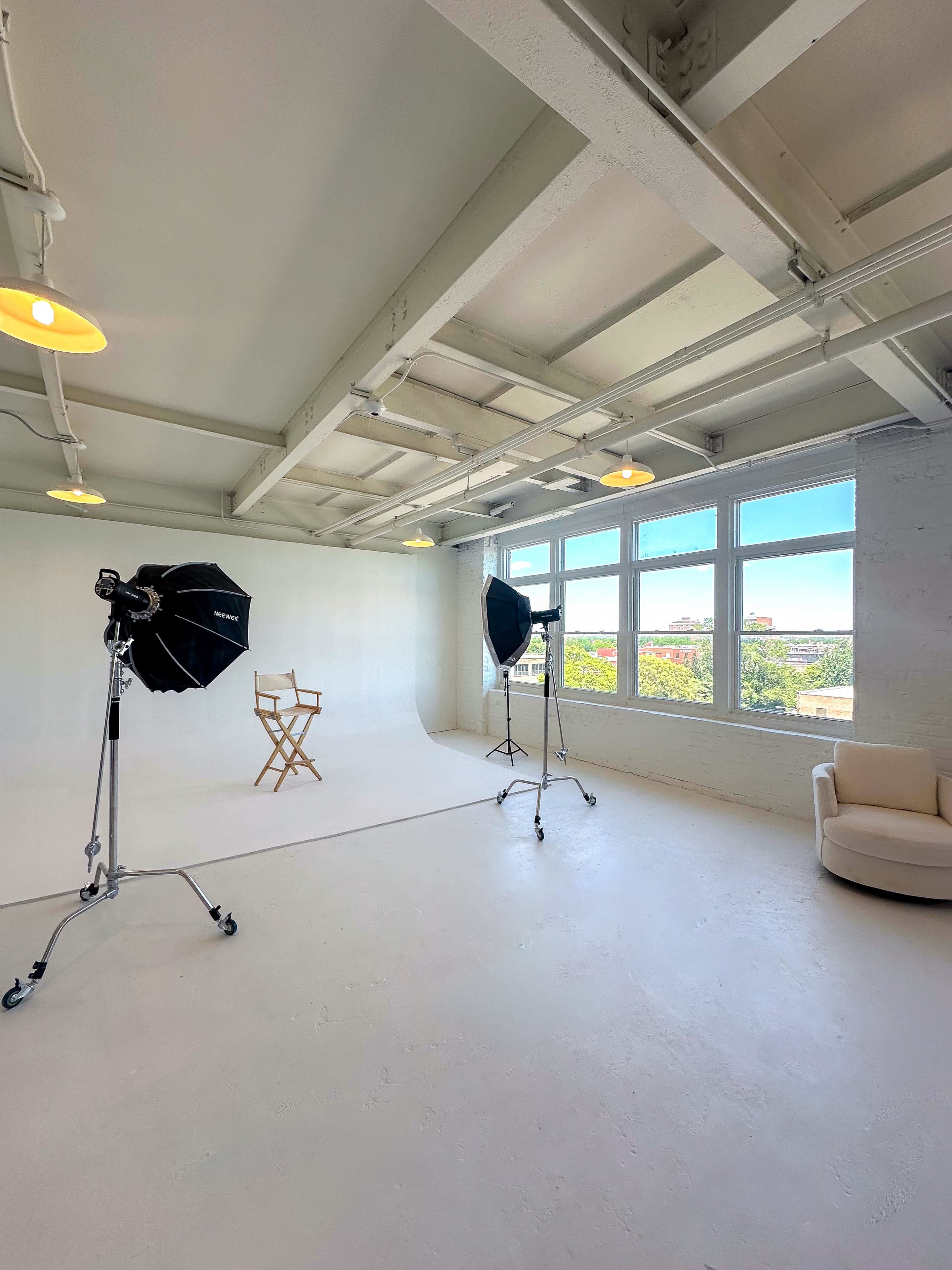 The image shows a well-lit photography studio with two softbox lights, a director's chair, and a beige armchair against a plain white backdrop.