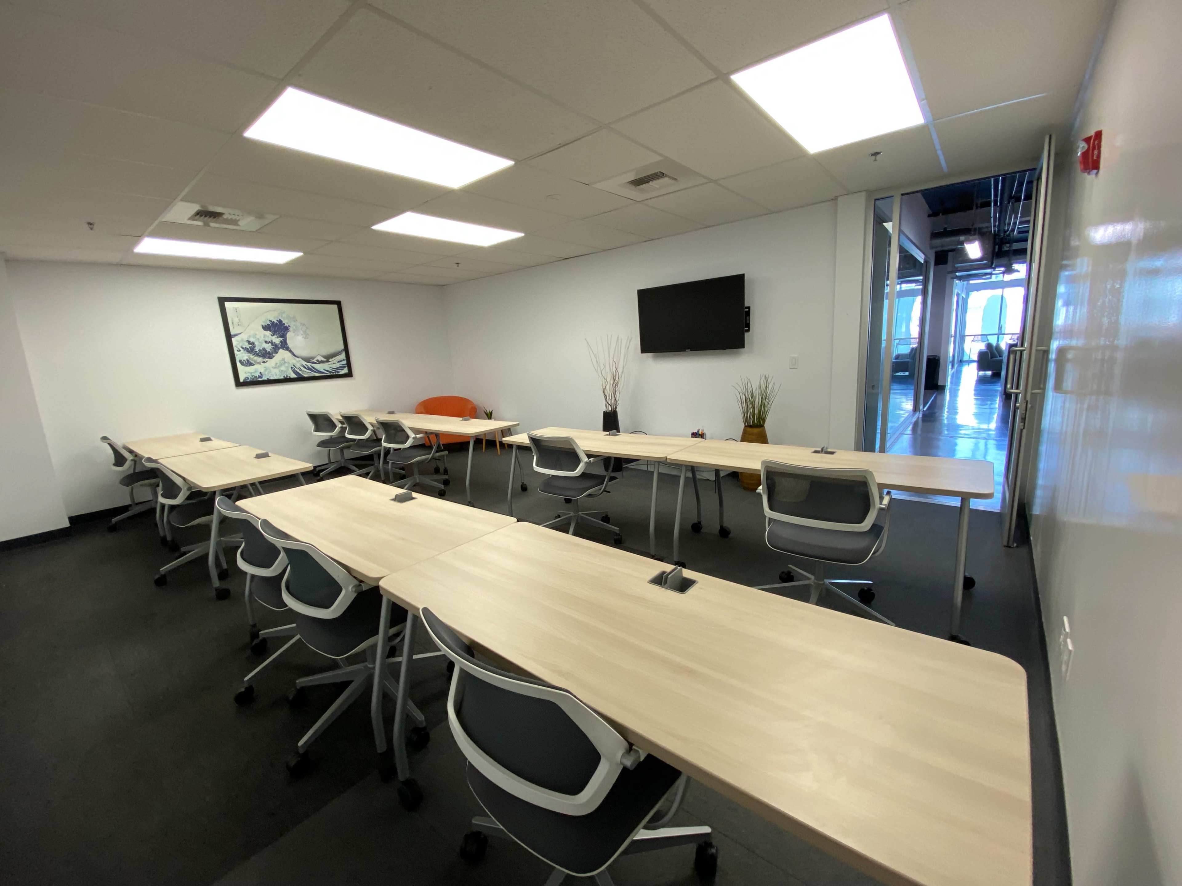 The image shows a modern conference room with multiple tables arranged in a U-shape, featuring a wall-mounted screen and minimalistic decor.
