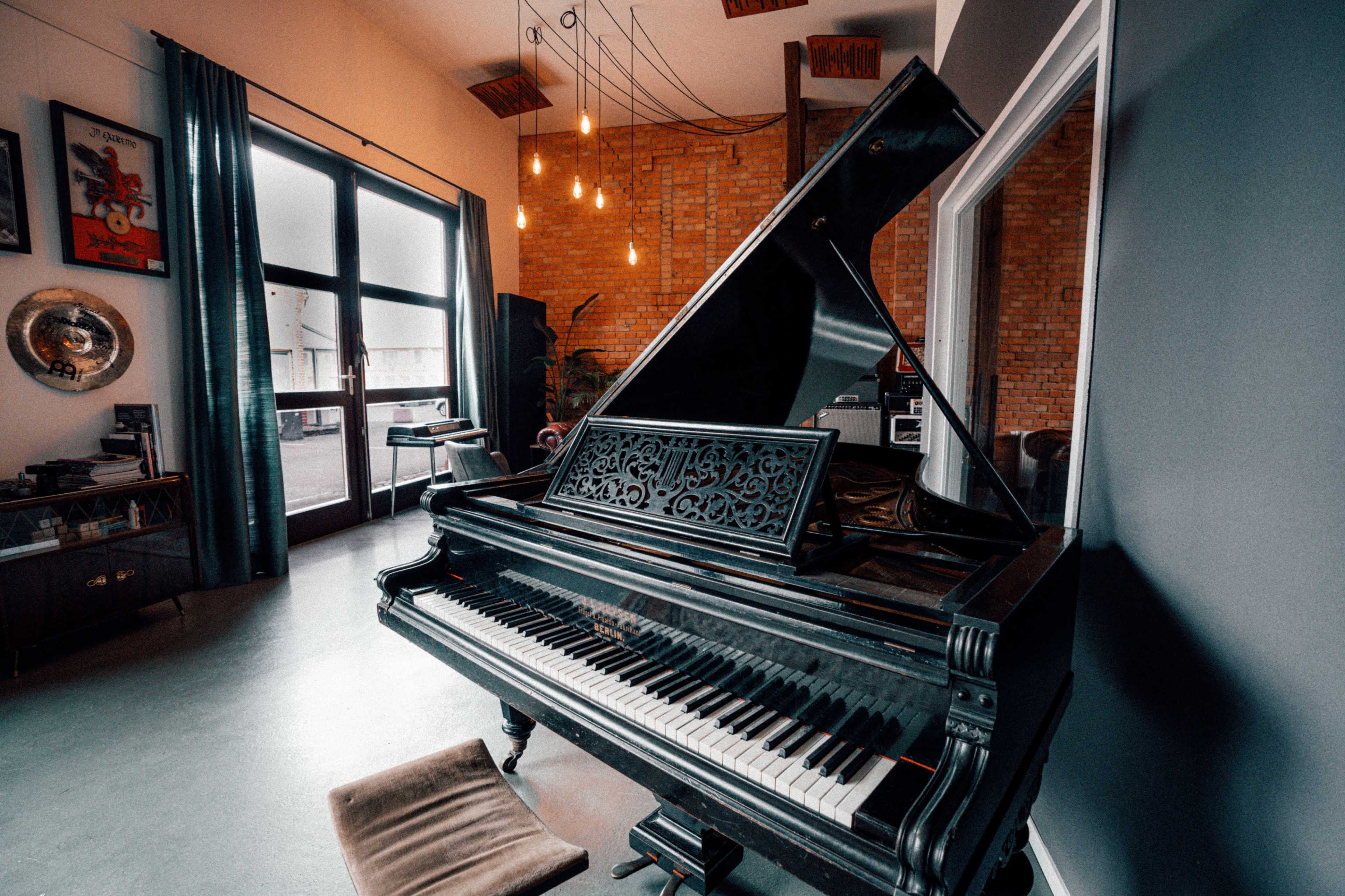 A grand piano sits in a modern living space with brick walls and large windows, illuminated by pendant lights.