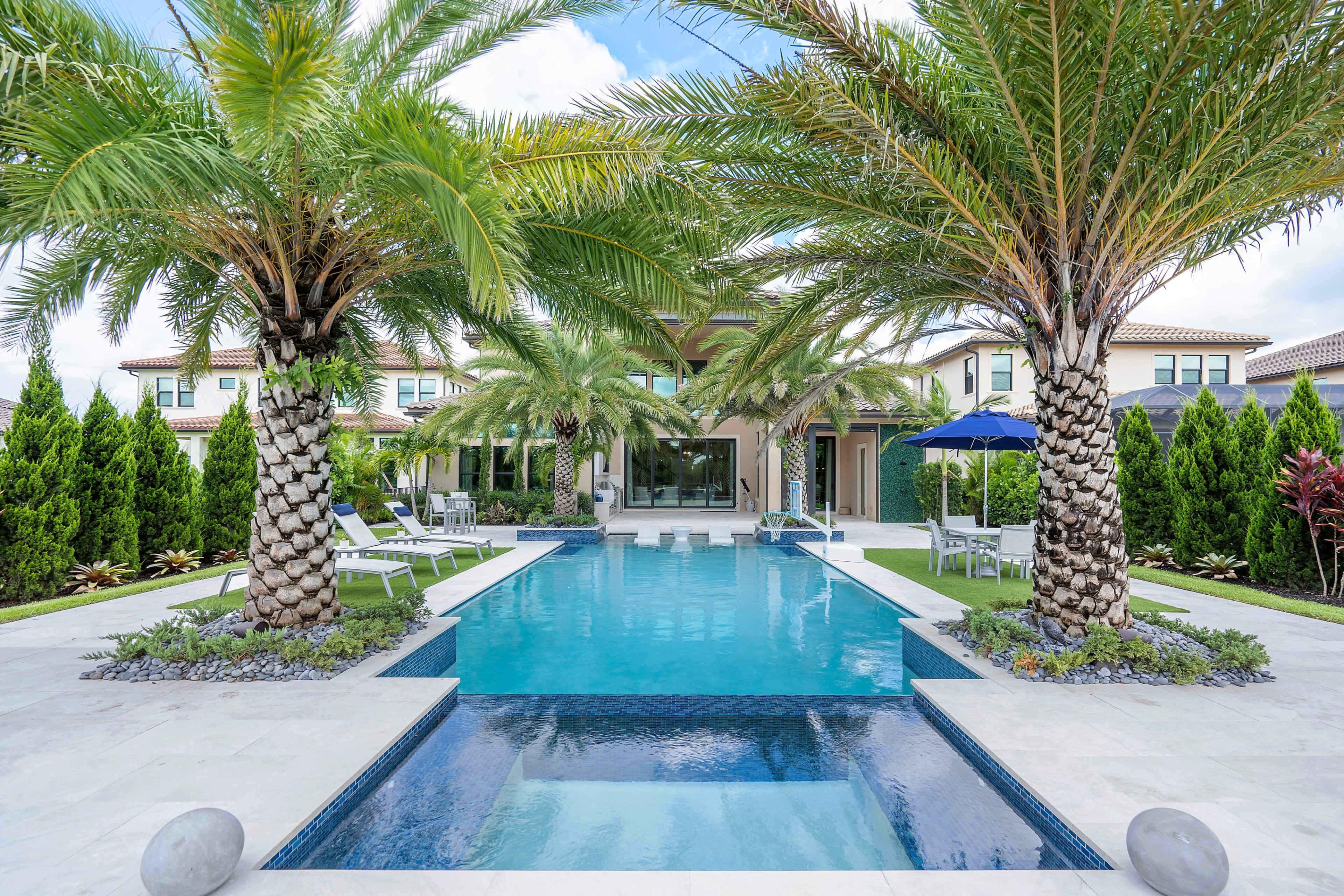 A modern backyard pool surrounded by palm trees, lounge chairs, and well-manicured greenery.