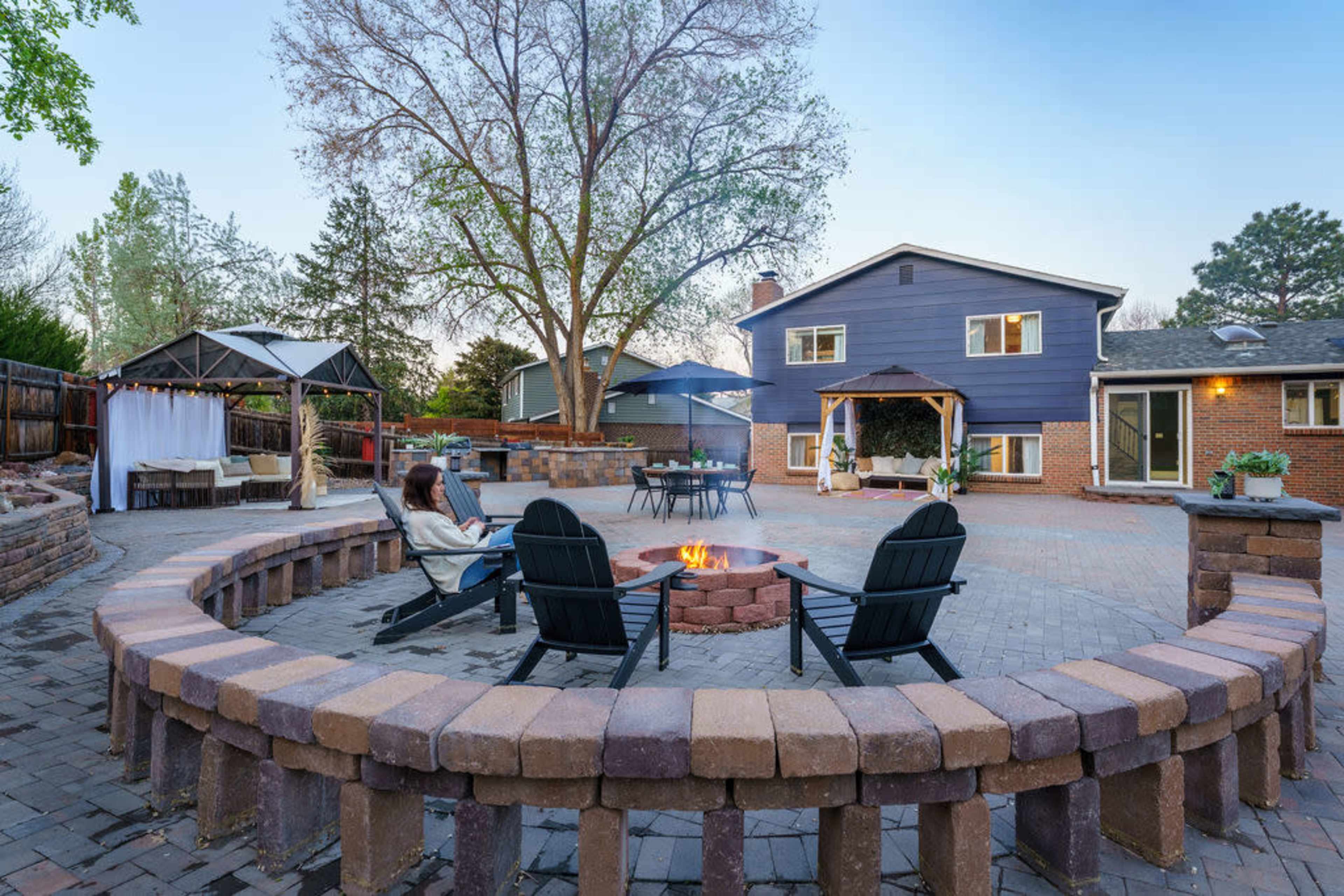 A circular stone fire pit surrounded by black adirondack chairs is located in a backyard with a two-story house and a gazebo visible in the background.