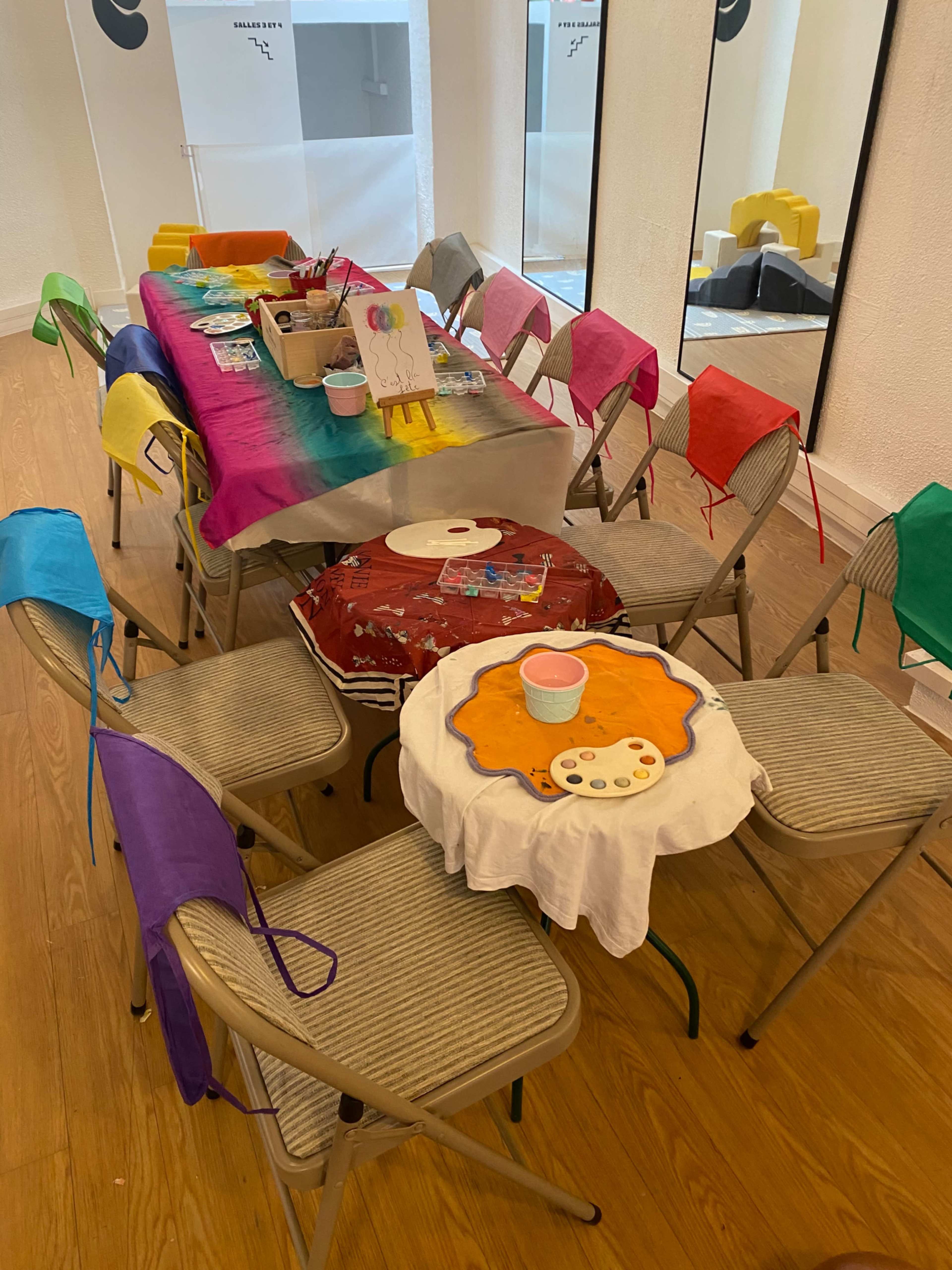 A colorful table set up for an art activity is surrounded by folding chairs, each adorned with a different colored apron.