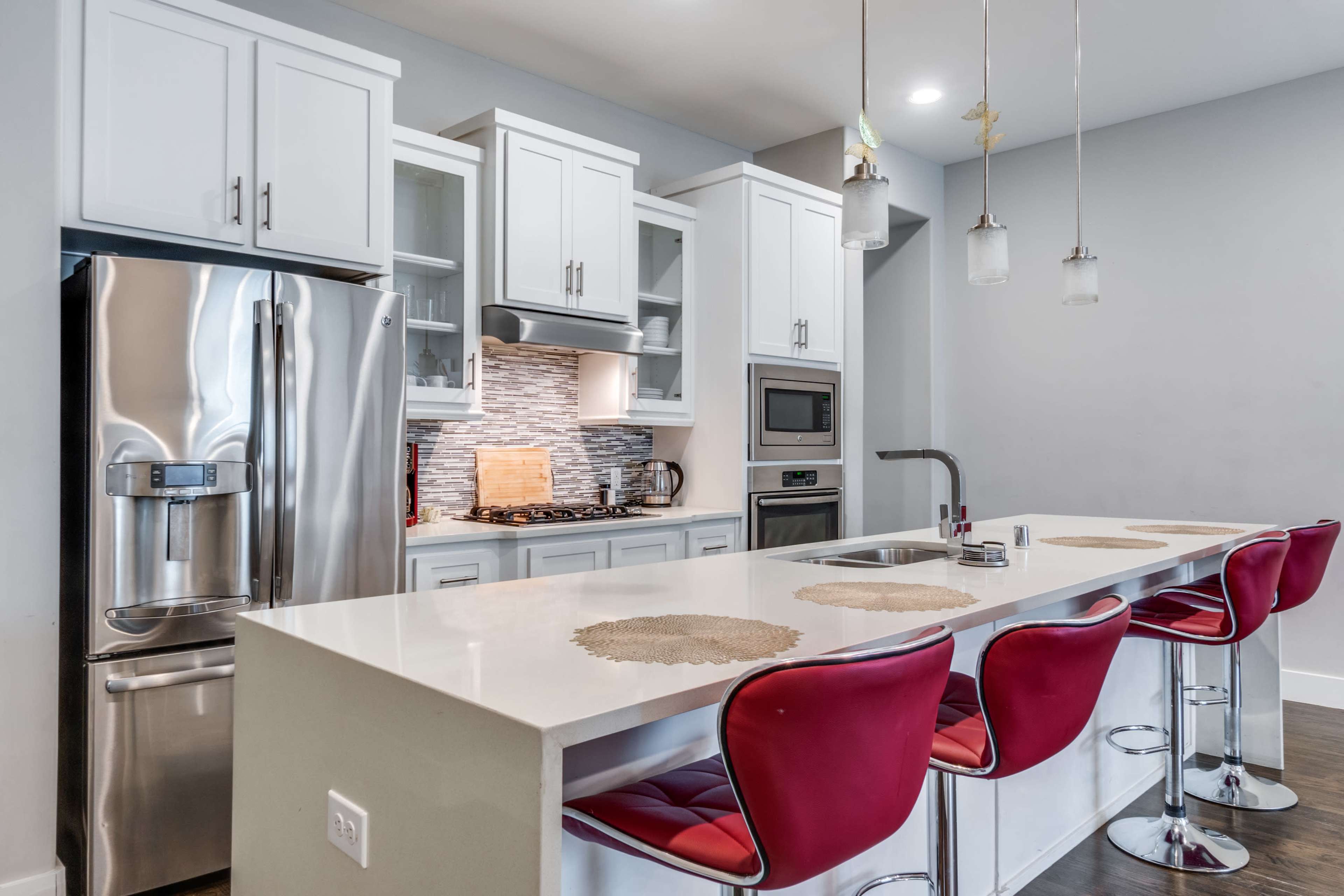 The image shows a modern kitchen with a large island, stainless steel appliances, and red bar stools.