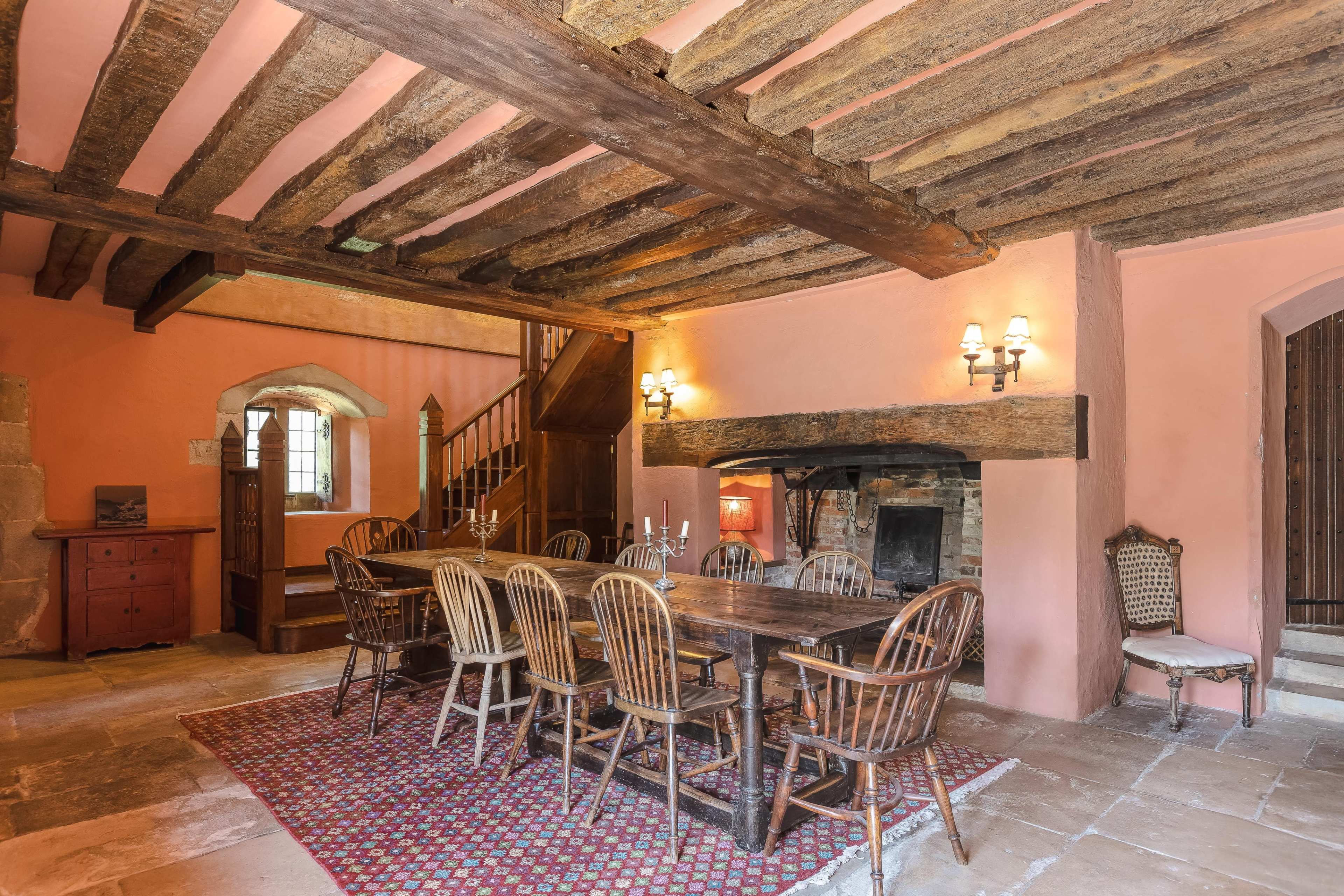 A rustic dining room featuring a long wooden table surrounded by chairs, with exposed beams on the ceiling and a stone fireplace.