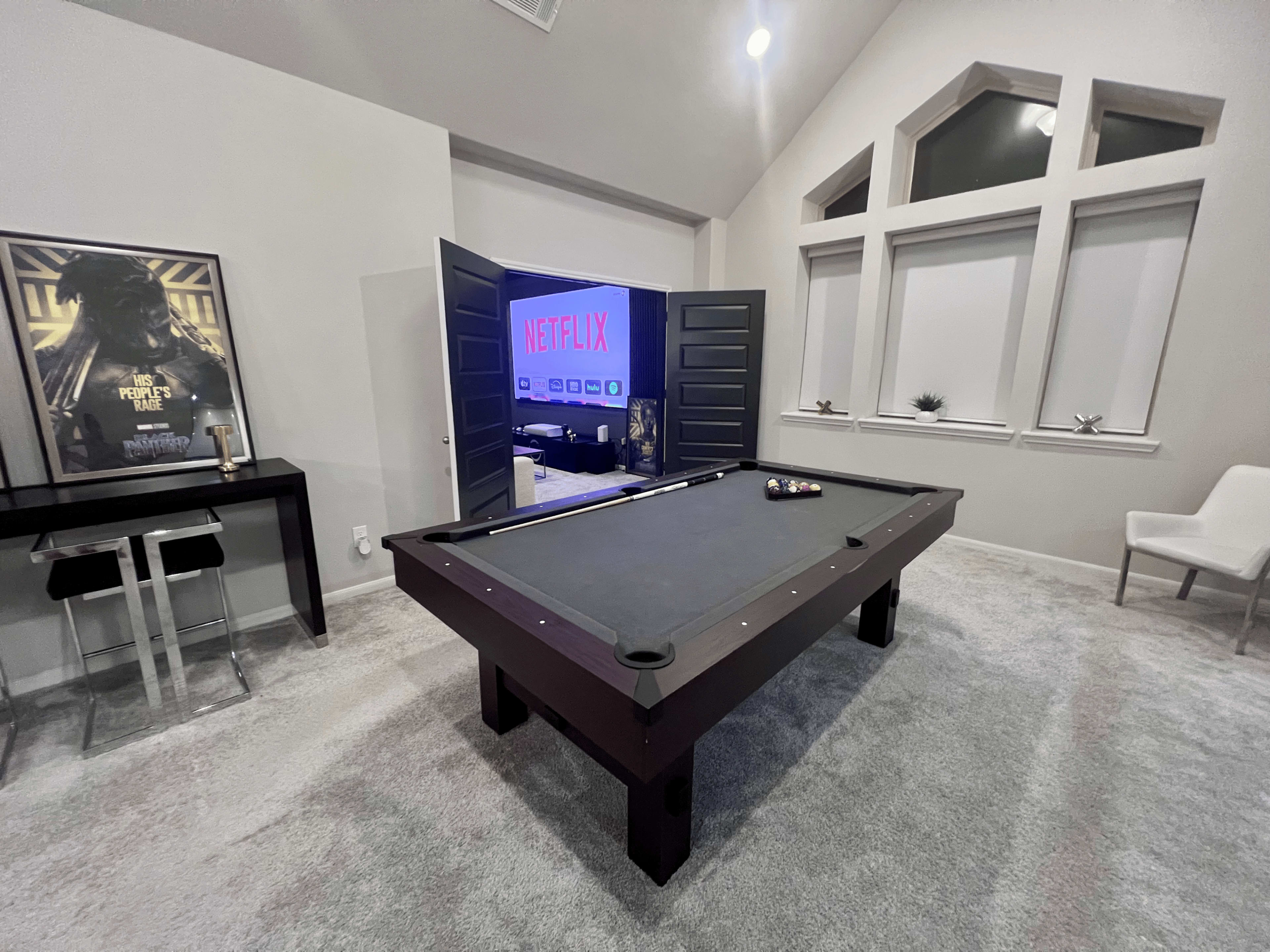 A modern room features a billiards table in the foreground, with an open doorway leading to a space illuminated by a Netflix sign in the background.