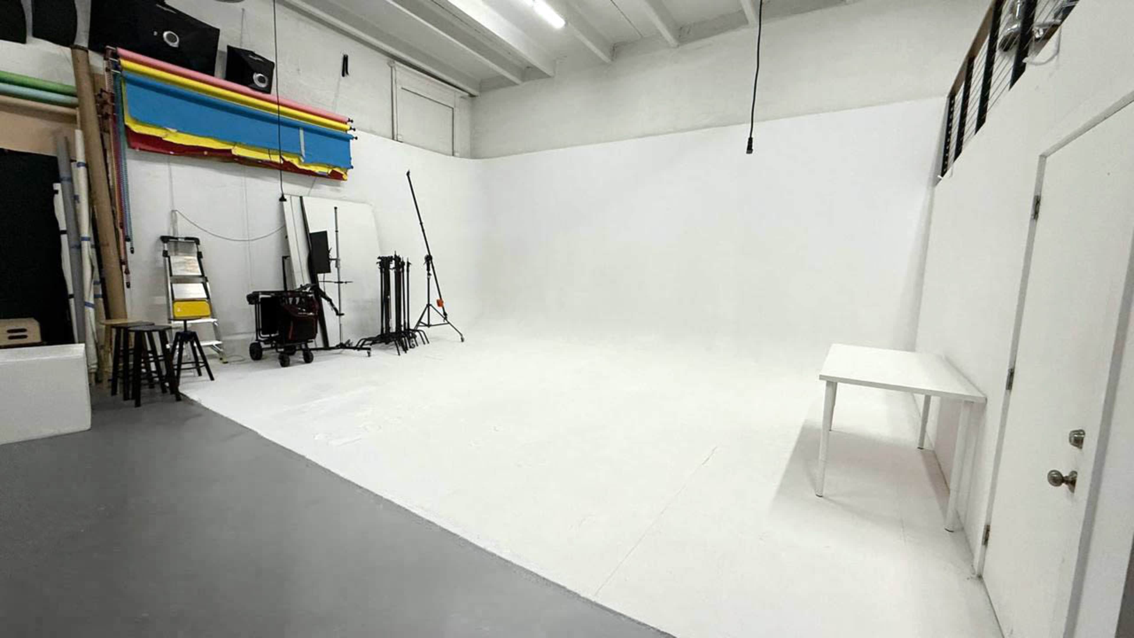 The image shows a spacious white photography studio with empty walls, a clean floor, and various equipment and props arranged along one side.