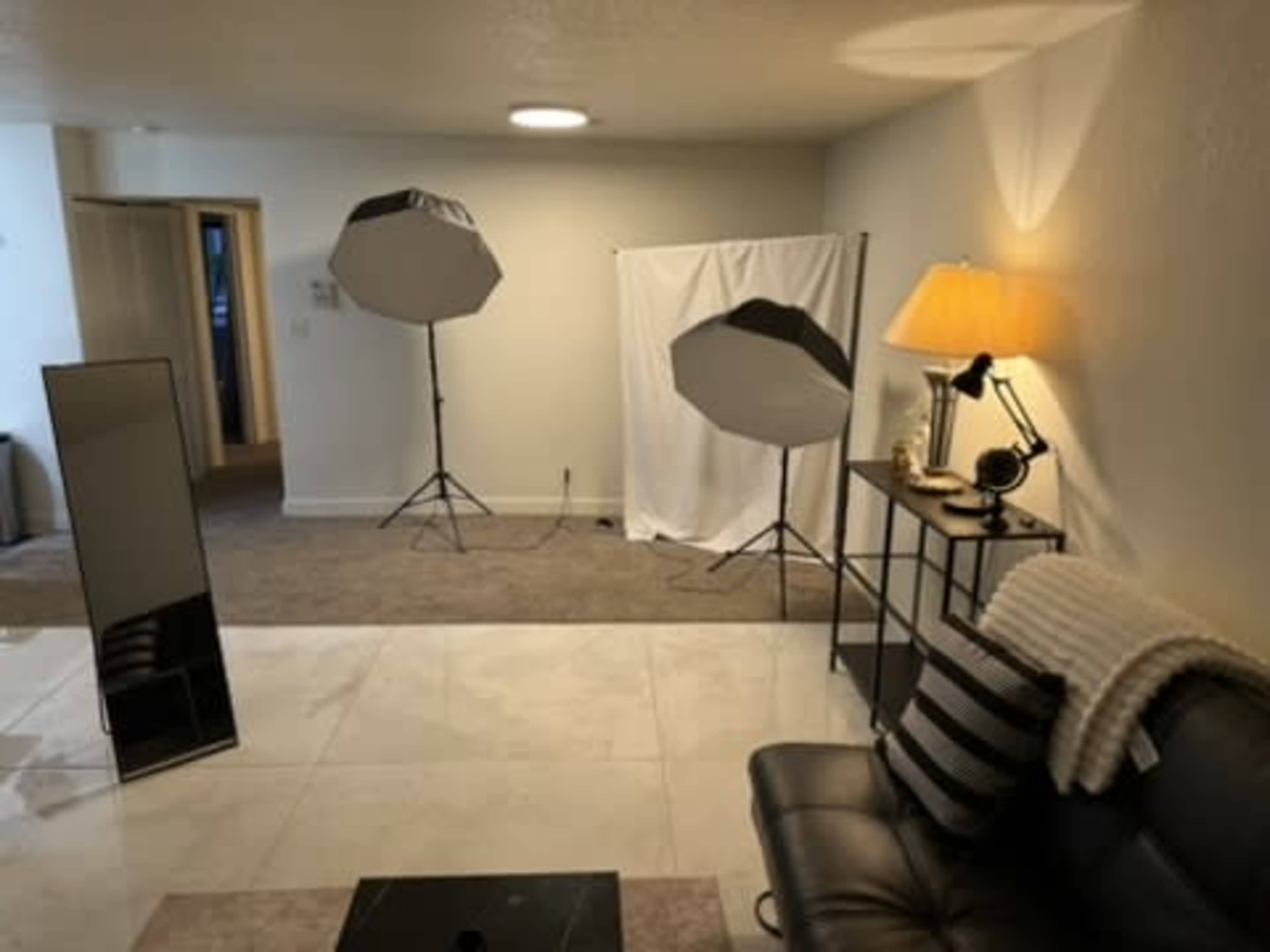 The image shows a well-lit room with a black sofa, a glass coffee table, a standing mirror, and two softbox lights set up near a white backdrop.