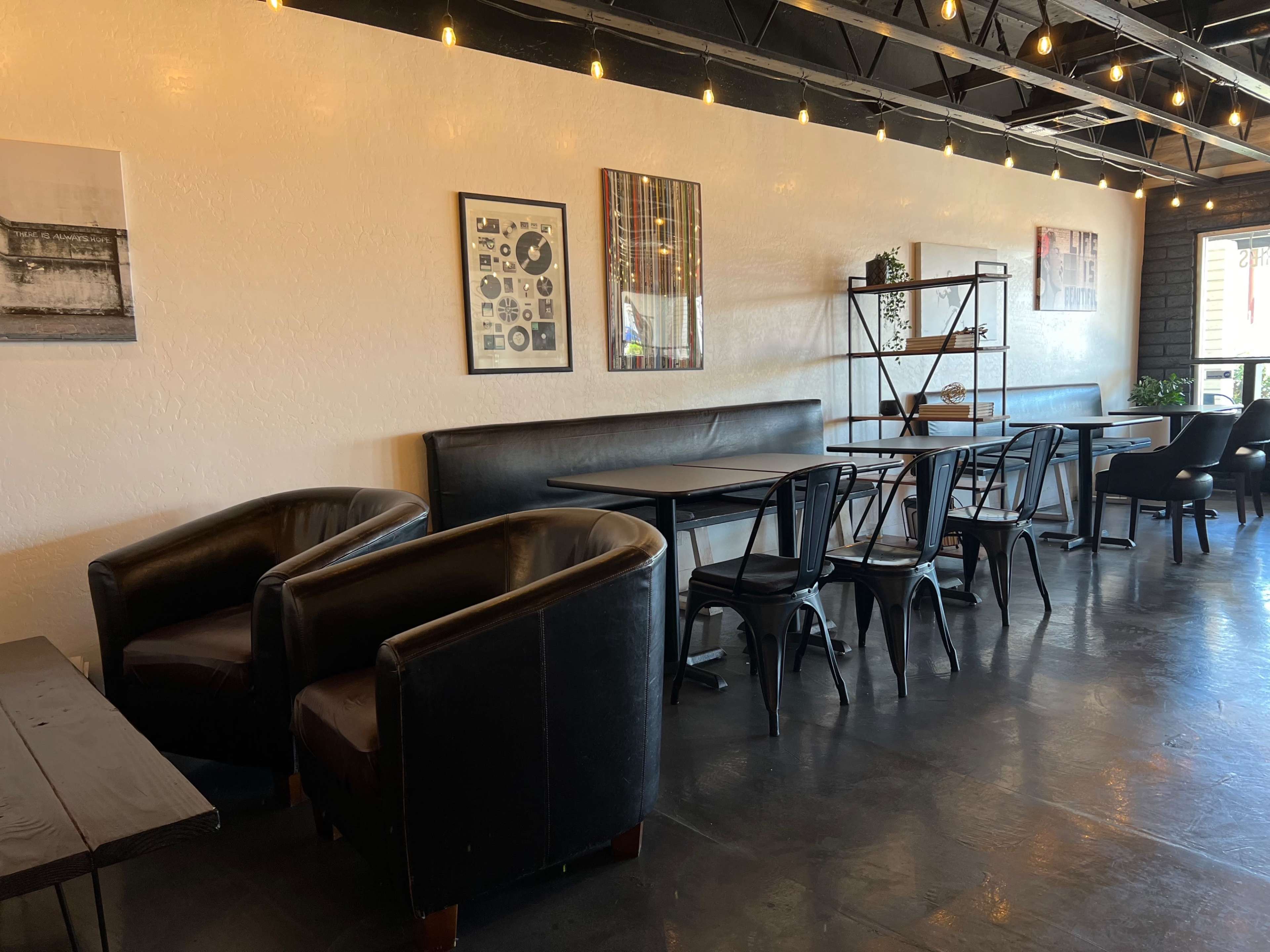 A cafe interior with black leather chairs, wooden tables, and framed artwork on the walls, illuminated by hanging string lights.
