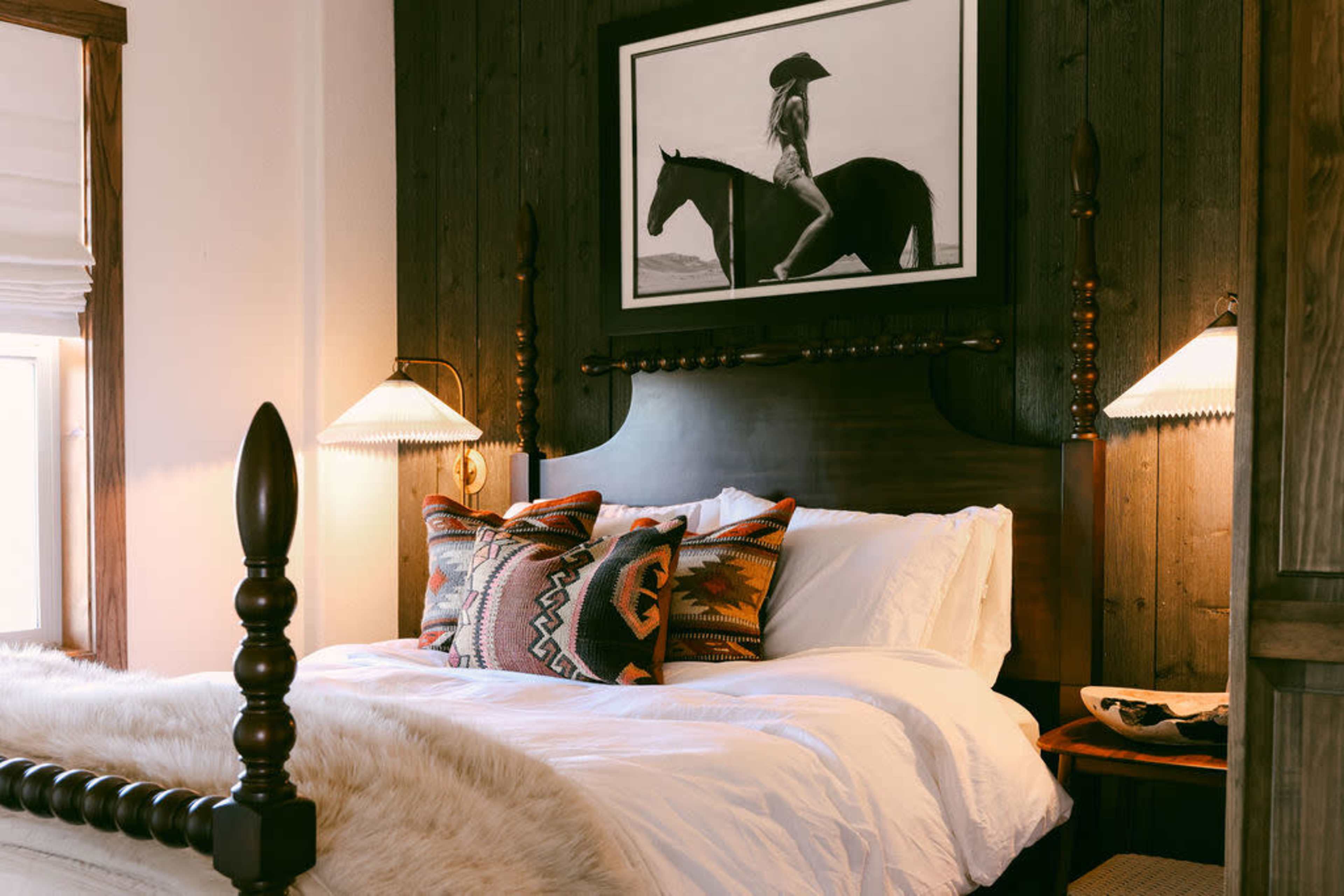 The image shows a cozy bedroom featuring a large bed with decorative pillows, a wooden headboard, and framed black-and-white artwork of a person on a horse hanging above it.