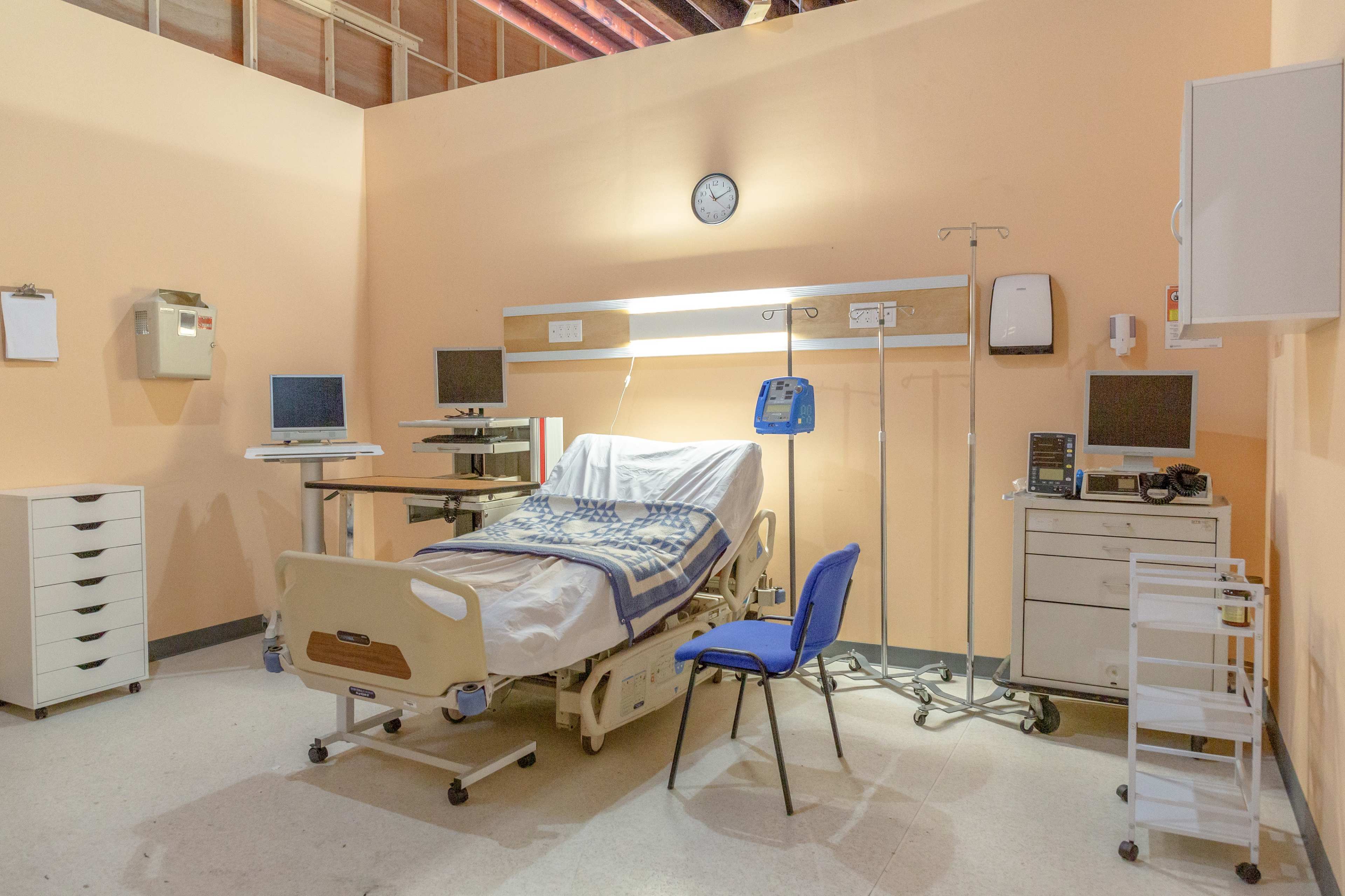 A hospital room featuring a bed with a blue and white checkered blanket, a chair, medical equipment, and a clock on the wall.