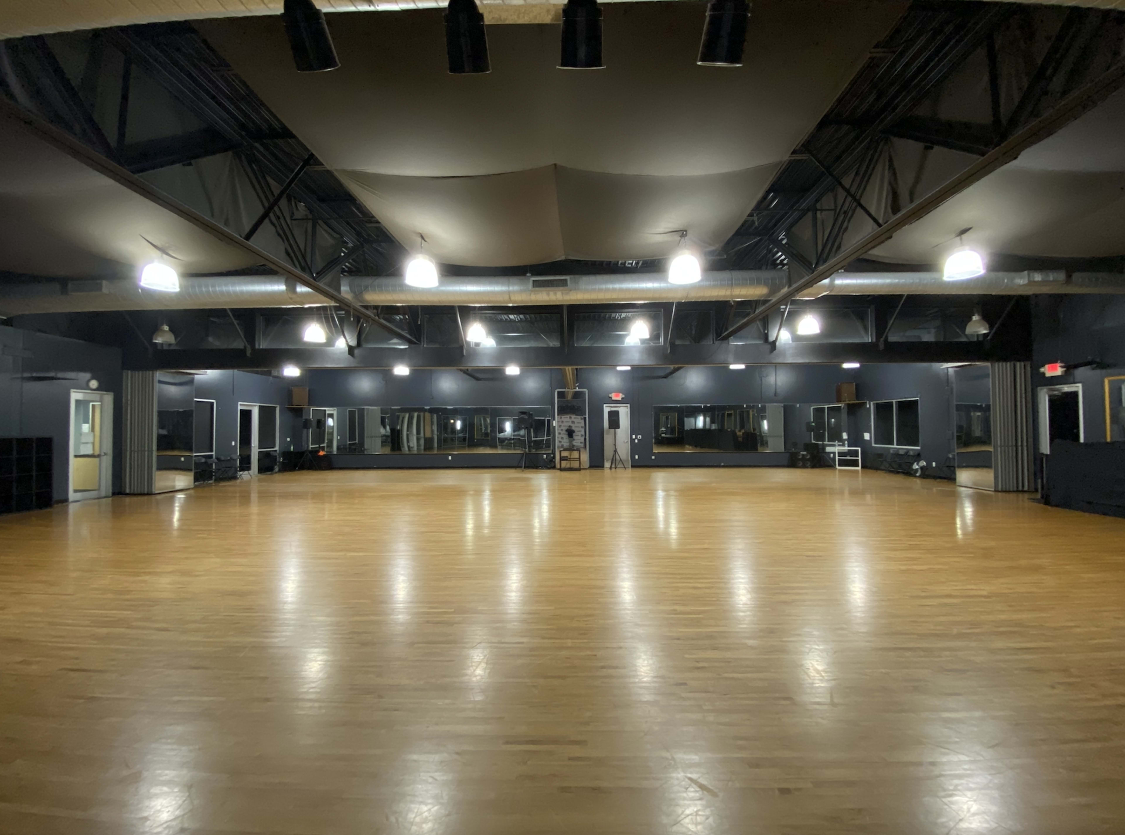 A large dance studio features a polished wooden floor, mirrored walls, and overhead lighting.