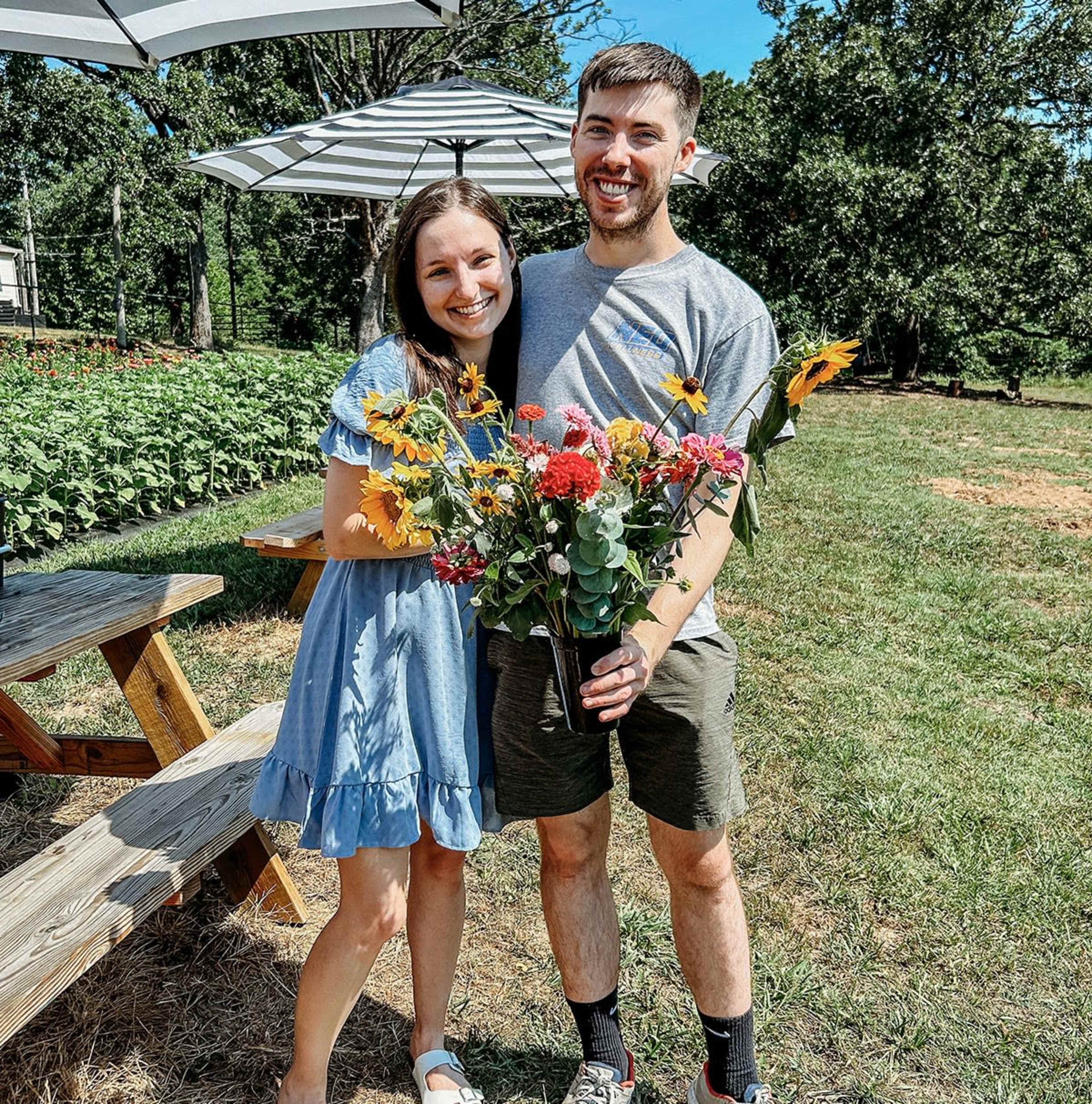 A couple stands smiling together in a field, holding a bouquet of colorful flowers.