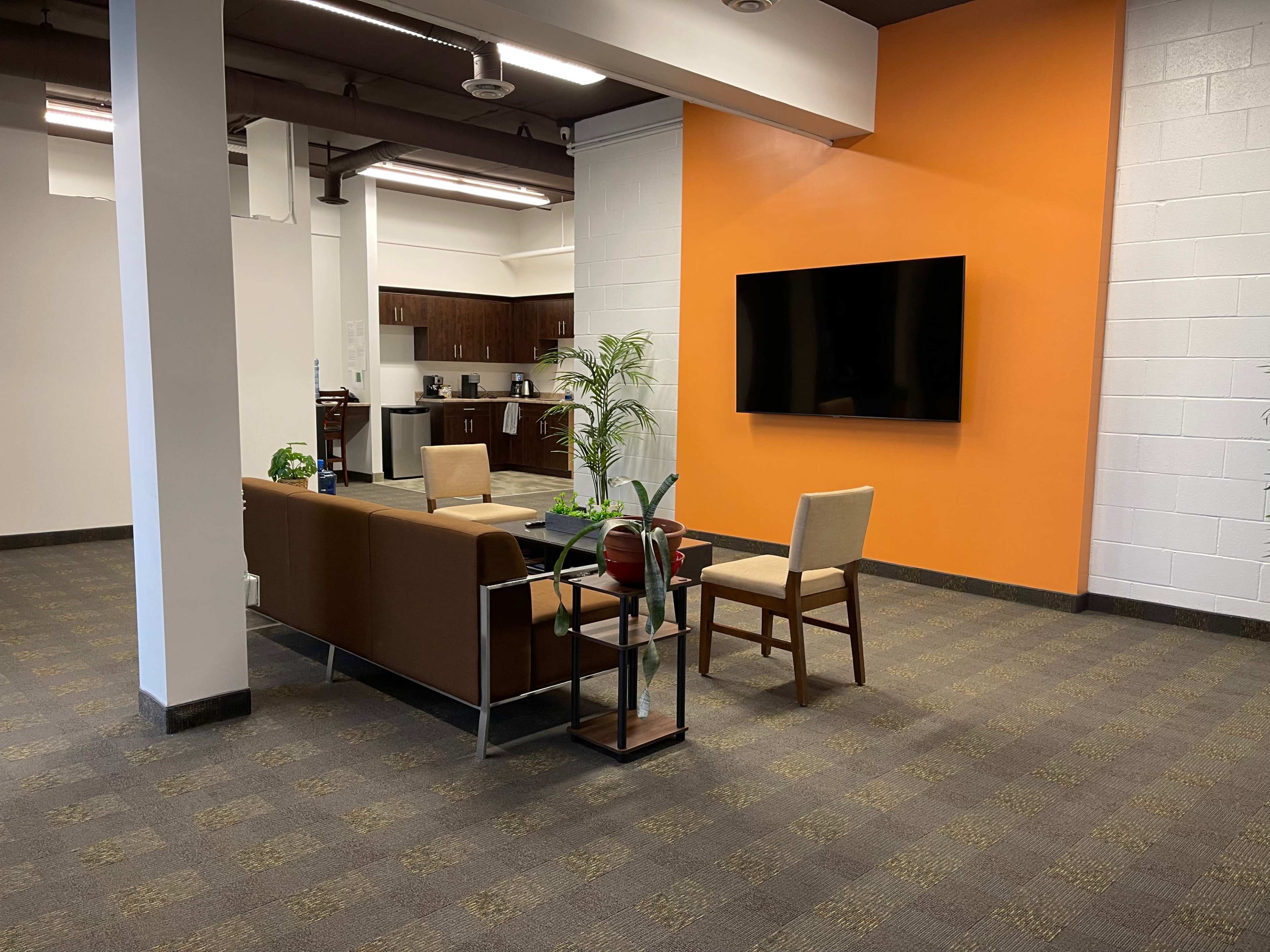 The image shows a modern lounge area with a brown sofa, a chair, a small table, and a wall-mounted television, set against an orange accent wall.