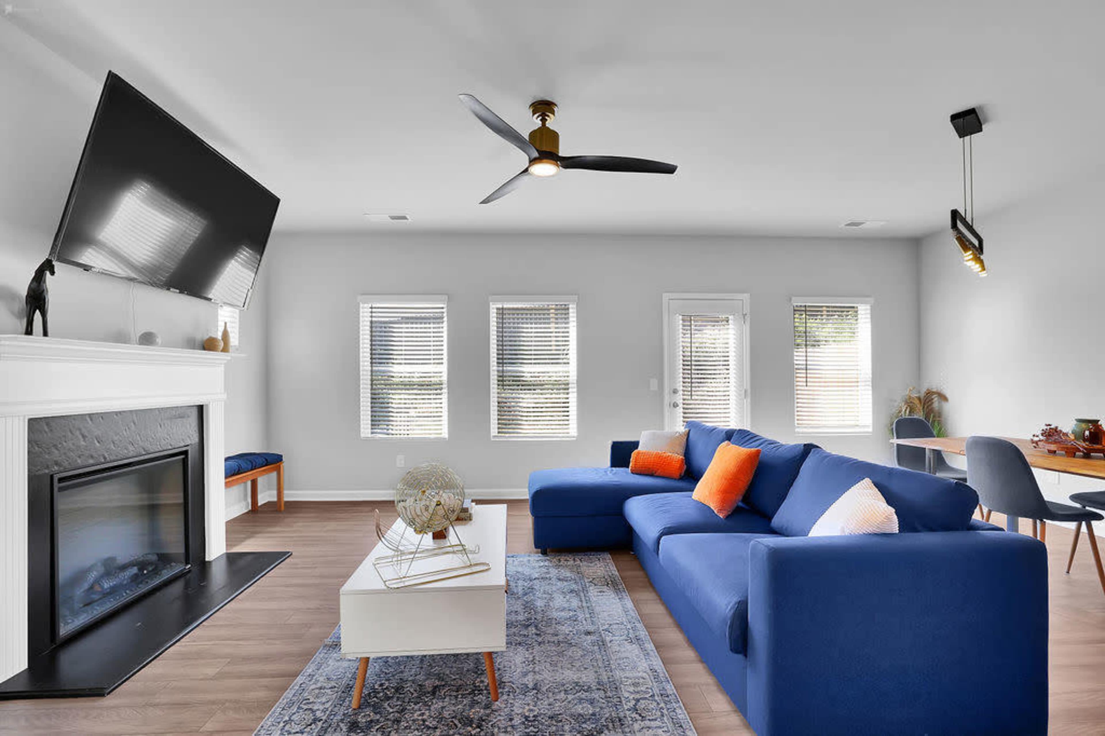 A modern living room with a blue sectional couch, a white coffee table, a large TV mounted above a fireplace, and several windows allowing natural light to enter.