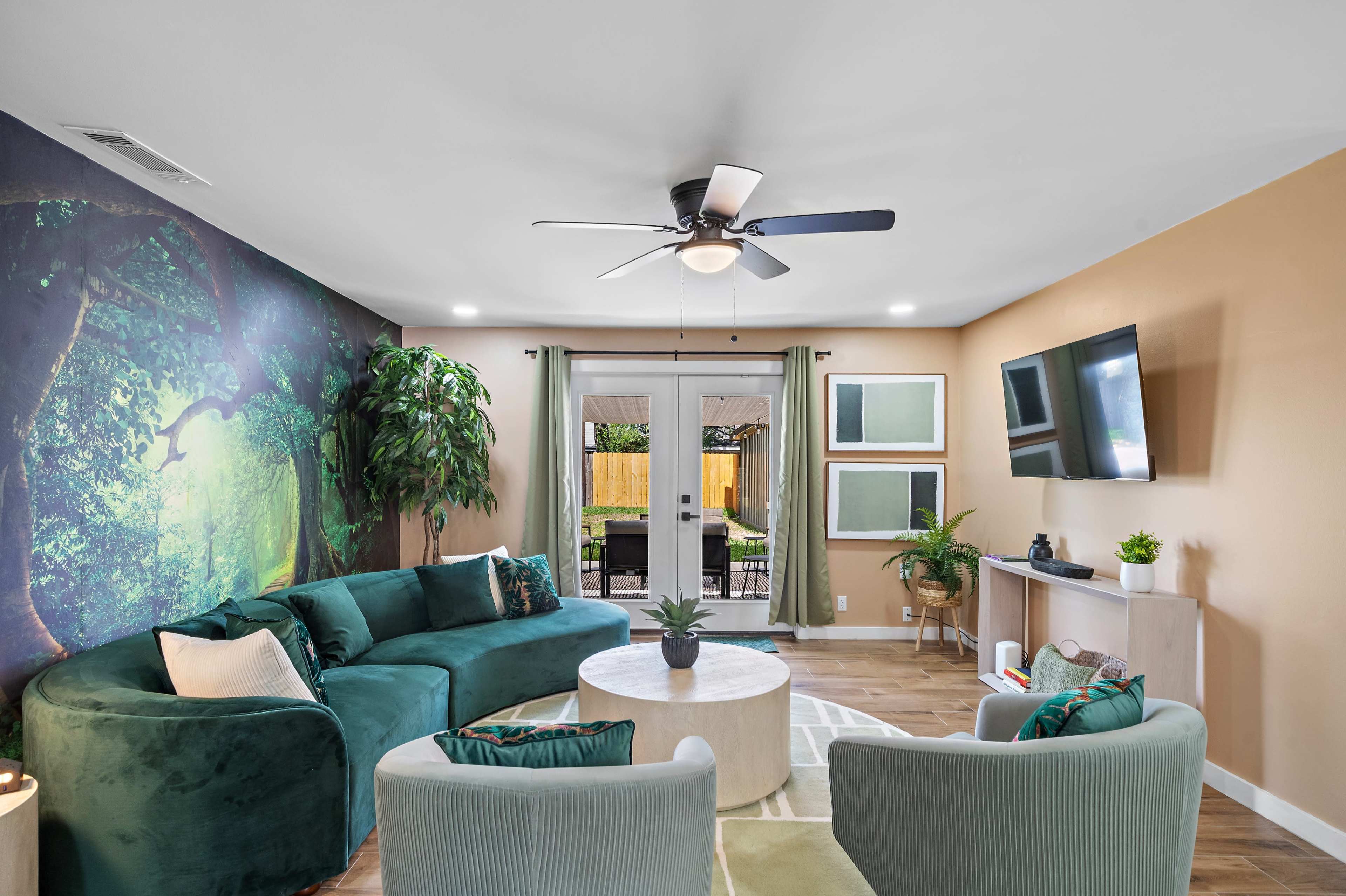 The living room features a green sectional sofa, a round coffee table, and large windows leading to a patio, adorned with a mural of trees on one wall.