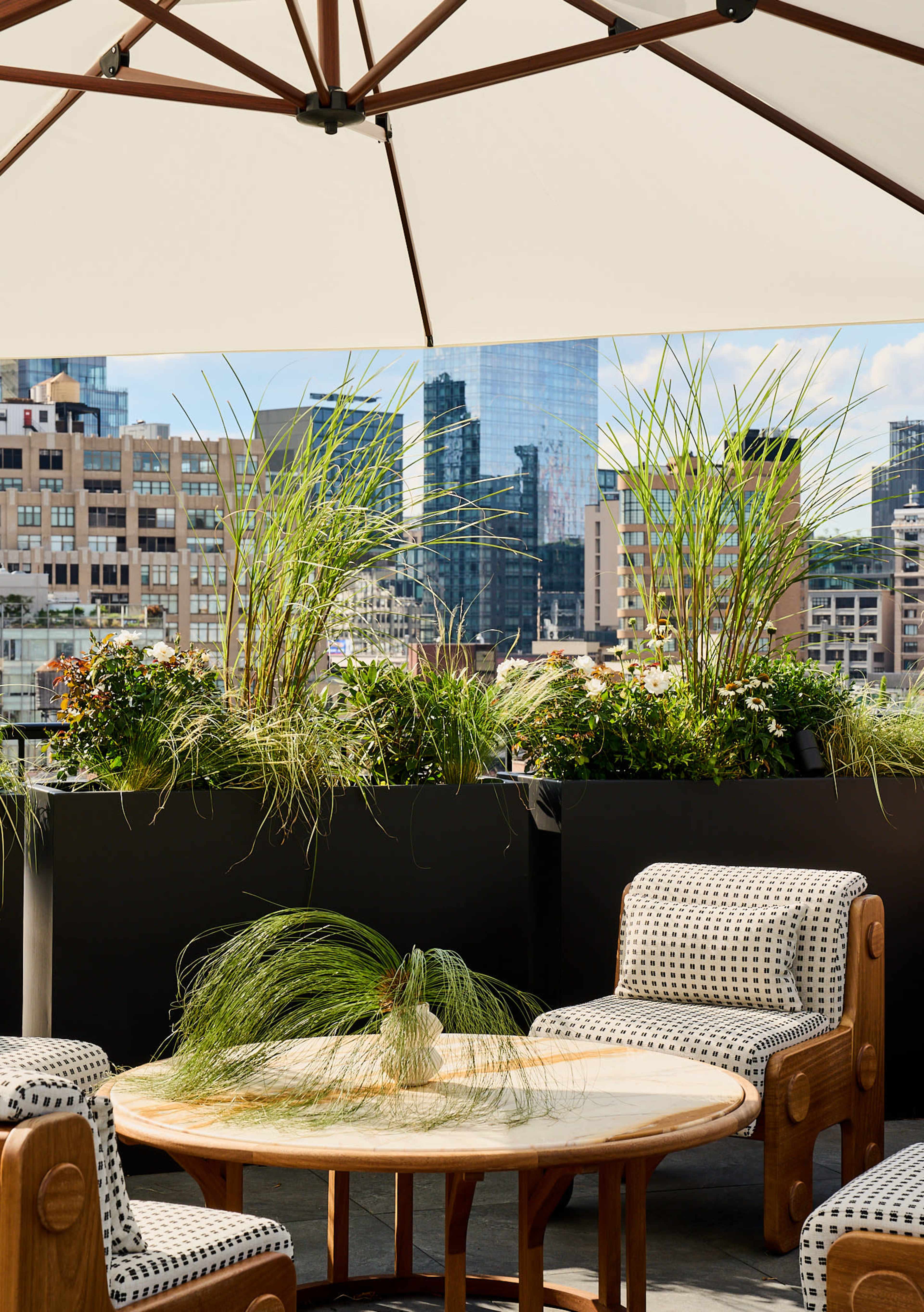 A rooftop terrace features a round wooden table surrounded by patterned chairs and planters with tall grasses, overlooking a city skyline.