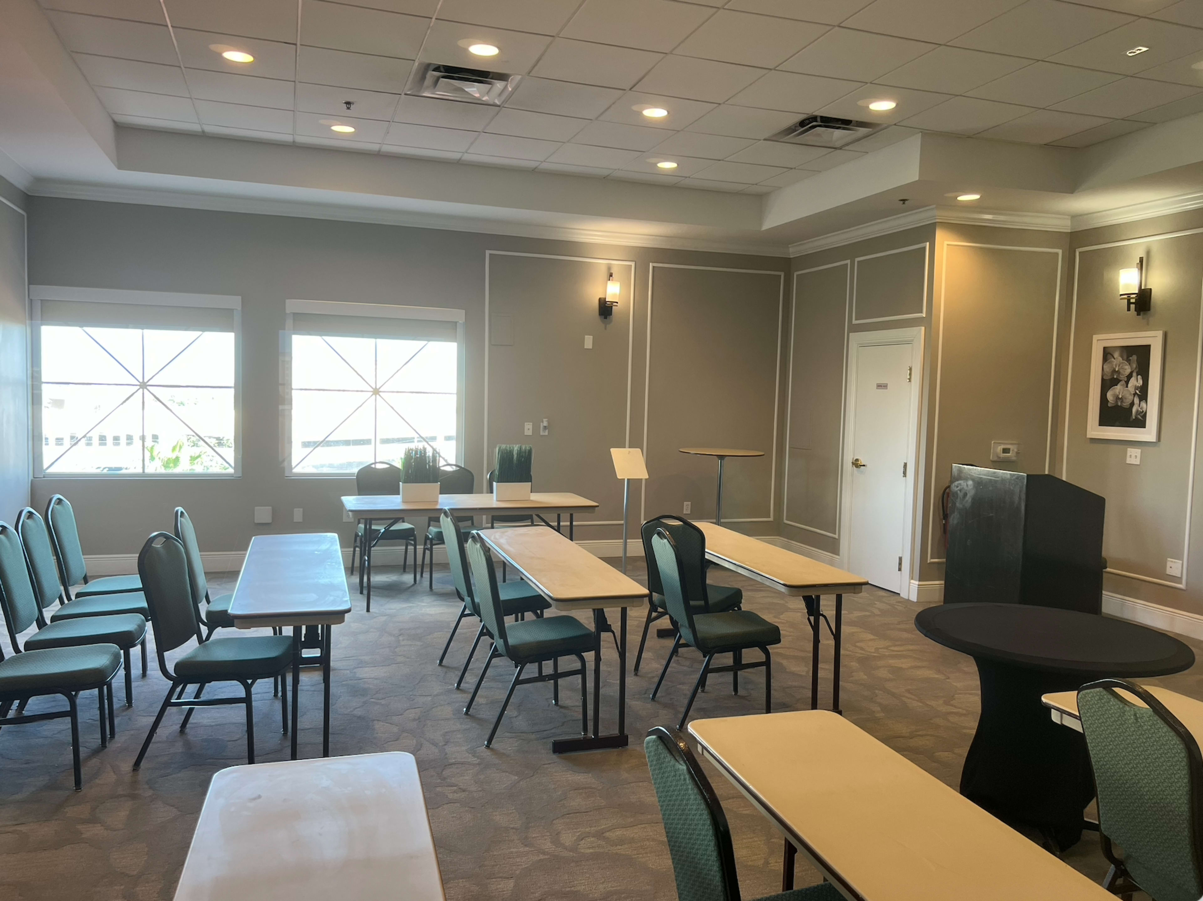 The image shows a meeting room with several rectangular tables and chairs arranged, a podium at the front, and large windows allowing natural light in.
