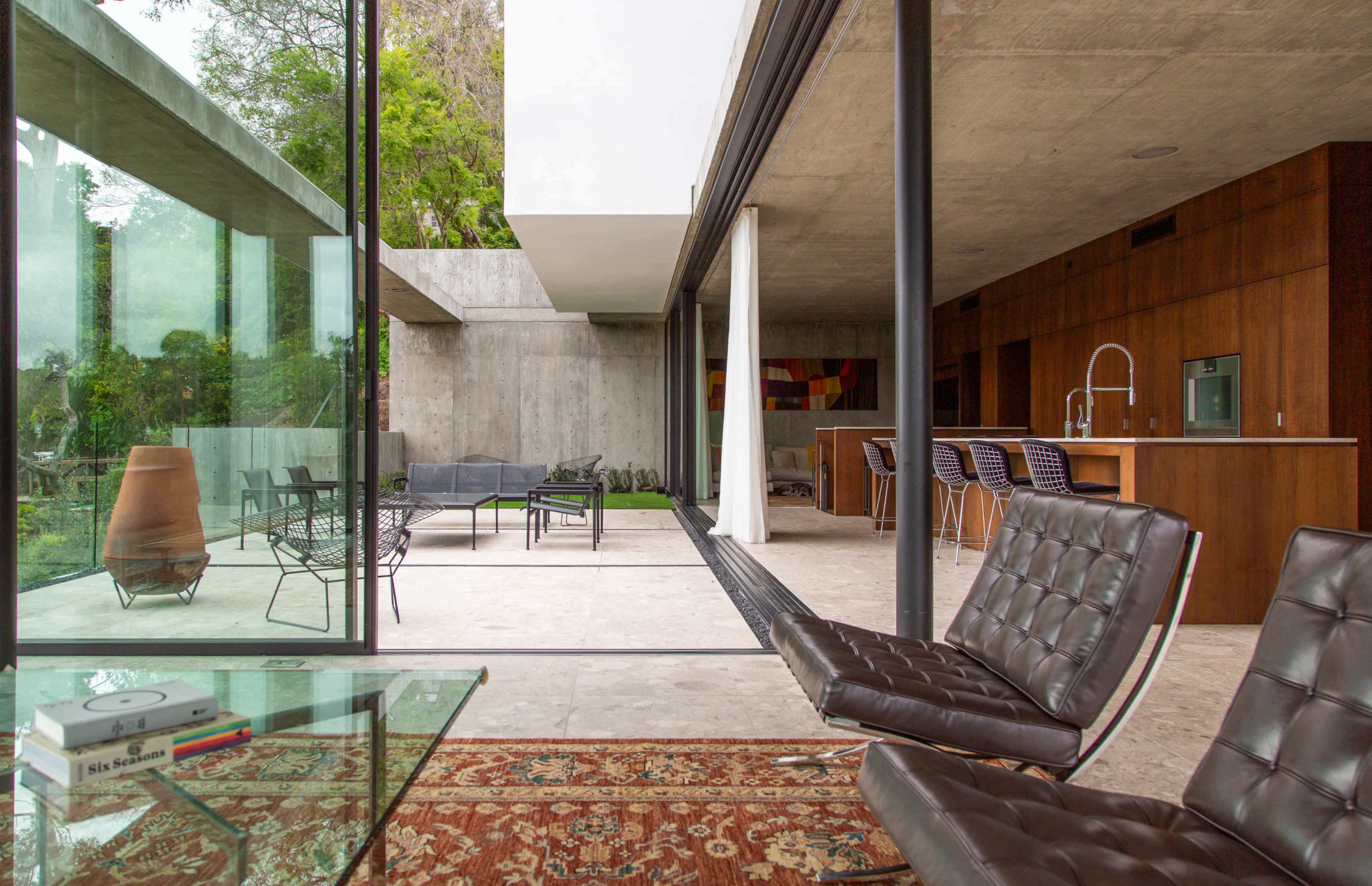 The image shows a modern indoor-outdoor living space featuring large glass walls, a concrete structure, minimalist furniture, and a compact garden area.