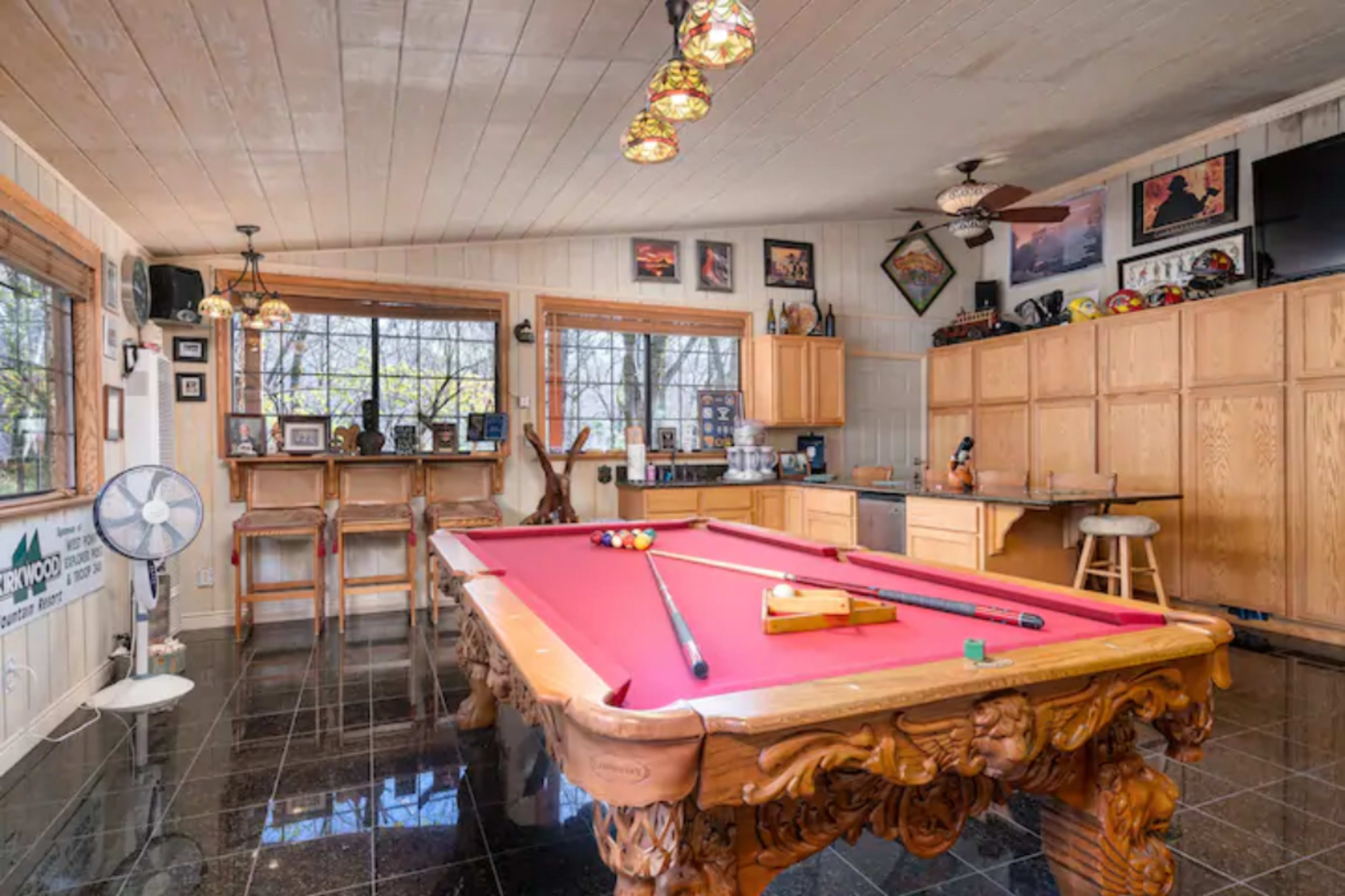 A room featuring a billiard table, wooden cabinets, and a bar area, all set against a backdrop of large windows.