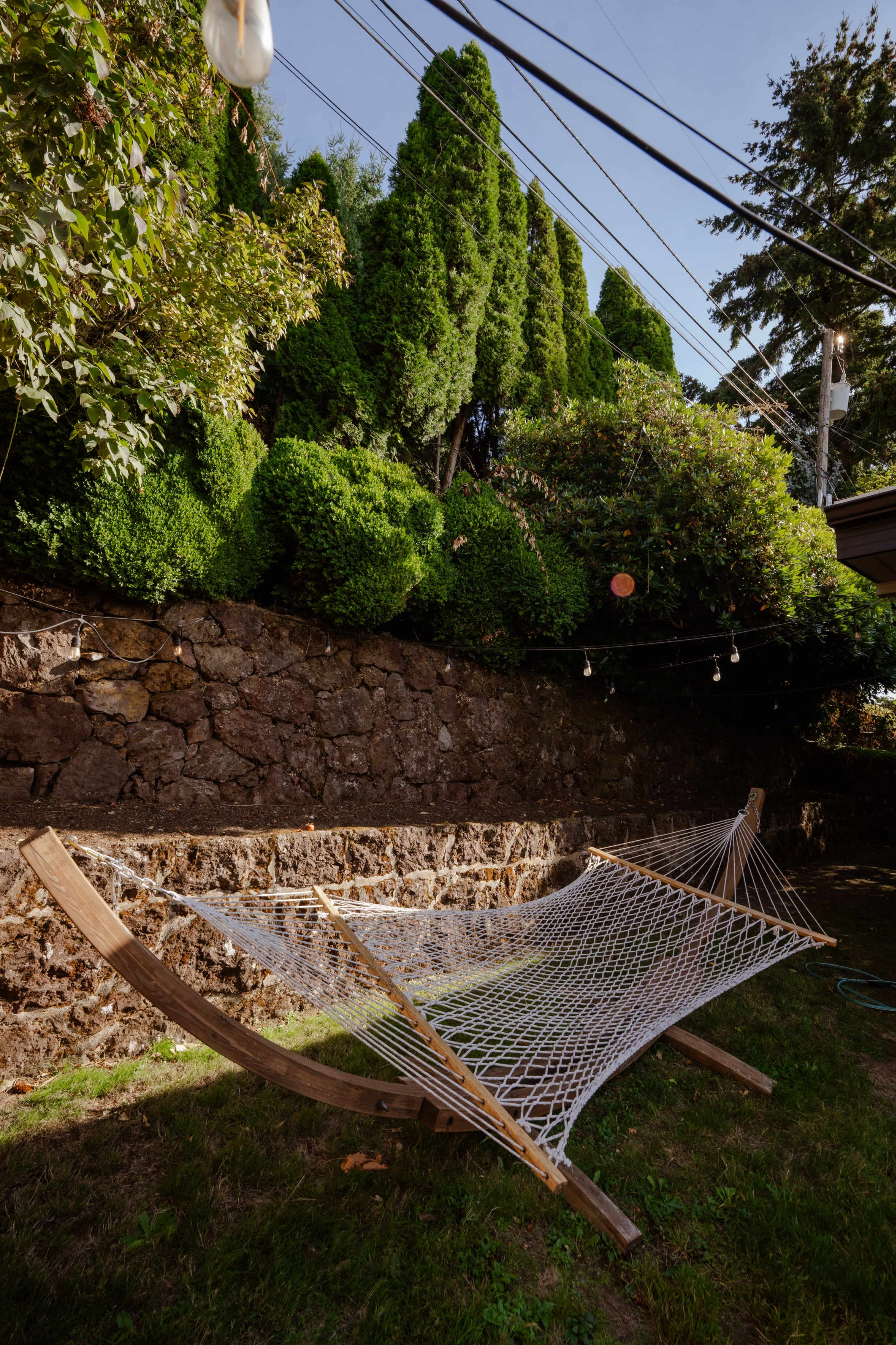 A hammock is situated in a grassy area against a stone wall, with lush greenery and trees above it.