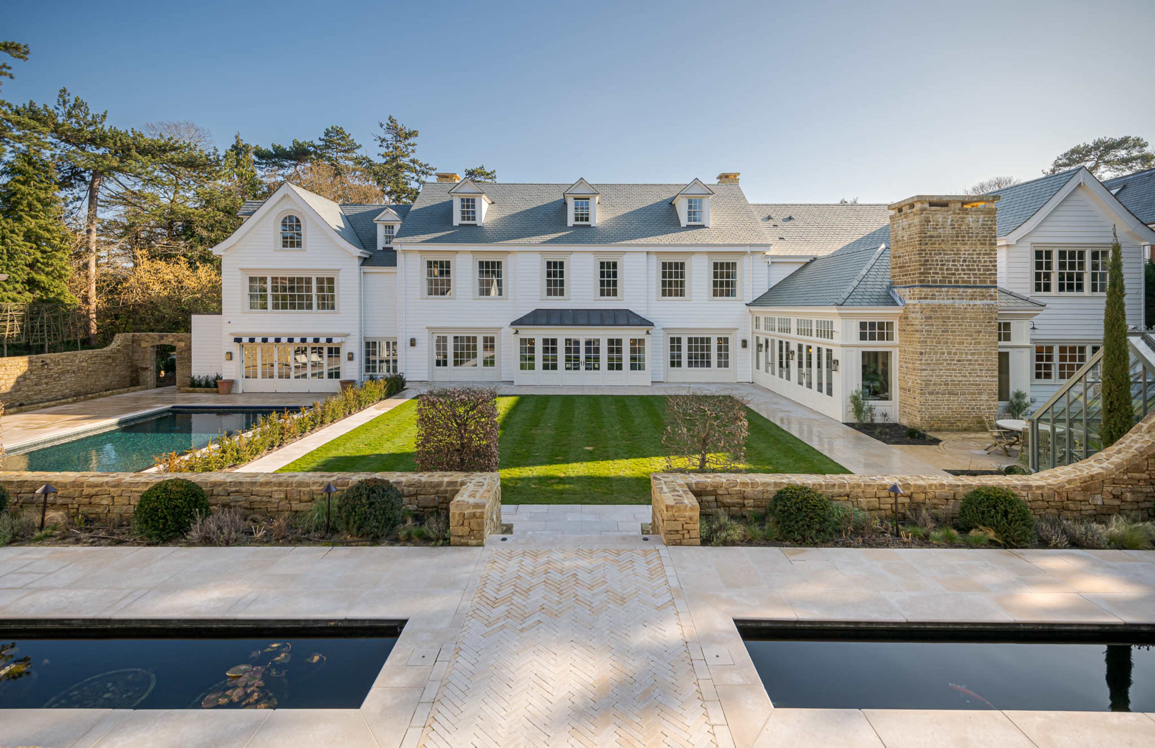A large, modern white house with multiple windows and an open courtyard features a swimming pool and landscaped gardens in the background.