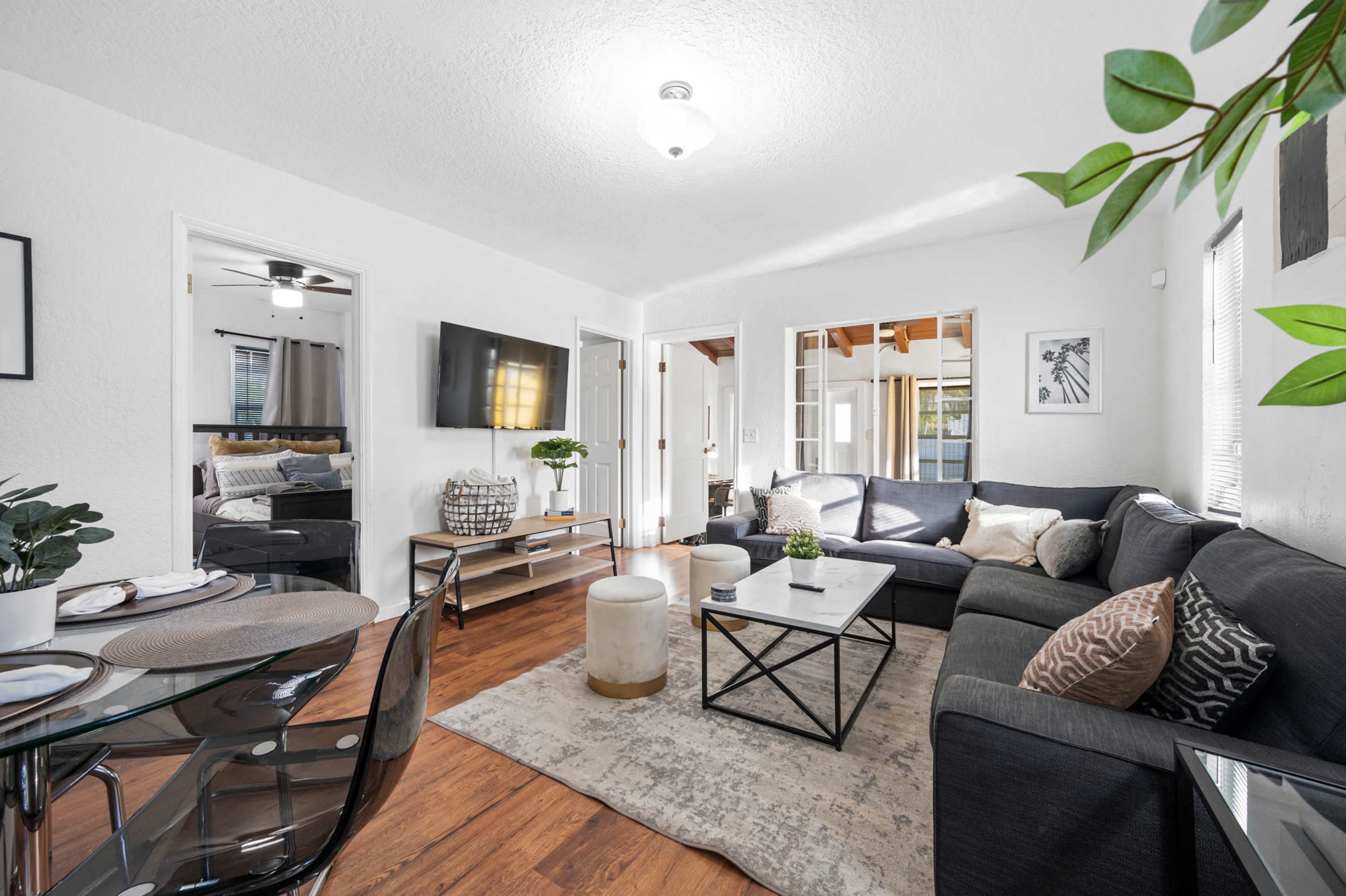 A modern living room features a dark sectional sofa, a coffee table, and a dining area with a glass table, accented by natural light and greenery.