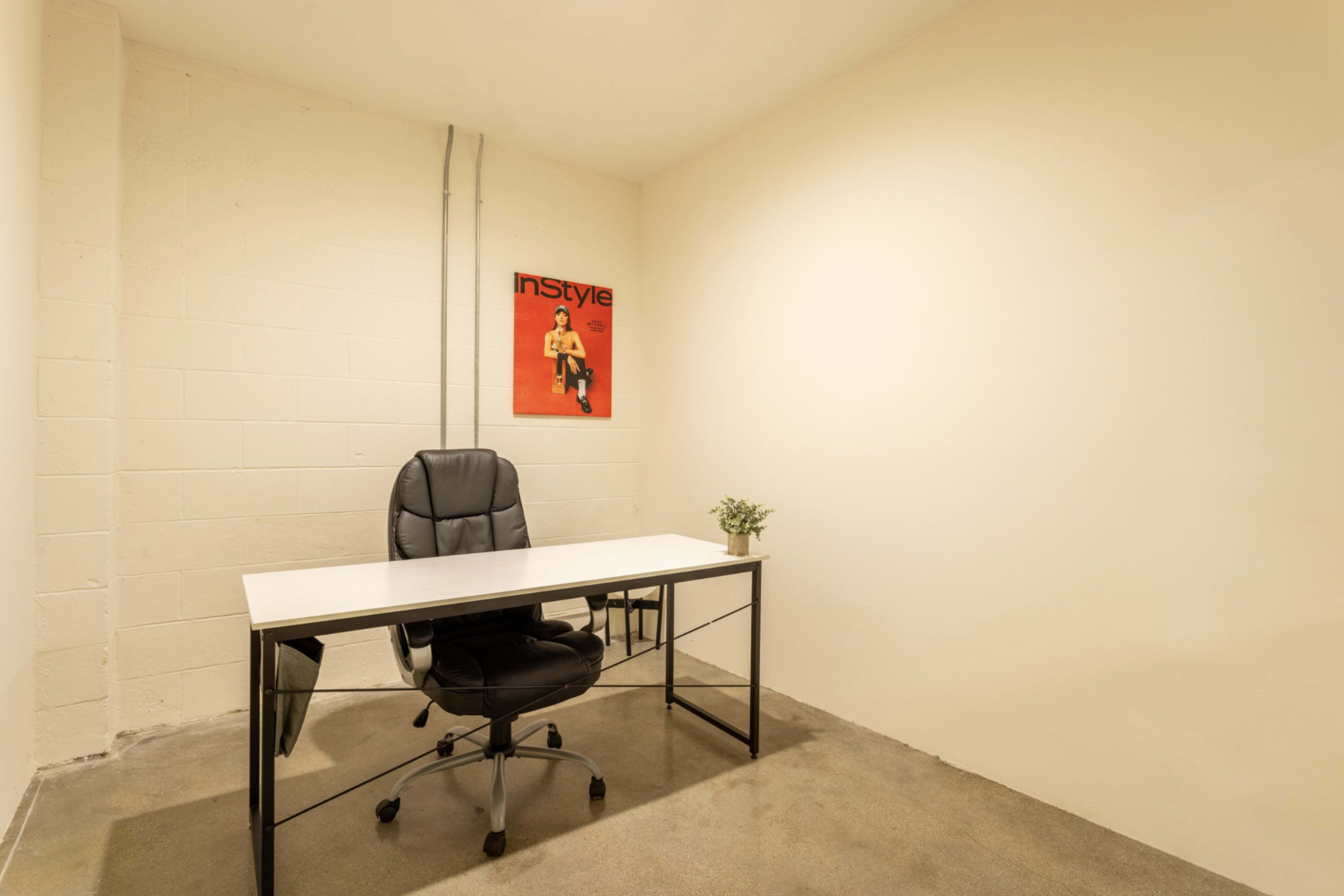 A minimalist office space featuring a black office chair and a white desk against a beige wall, with a framed poster hanging above the desk and a small potted plant on the desk.