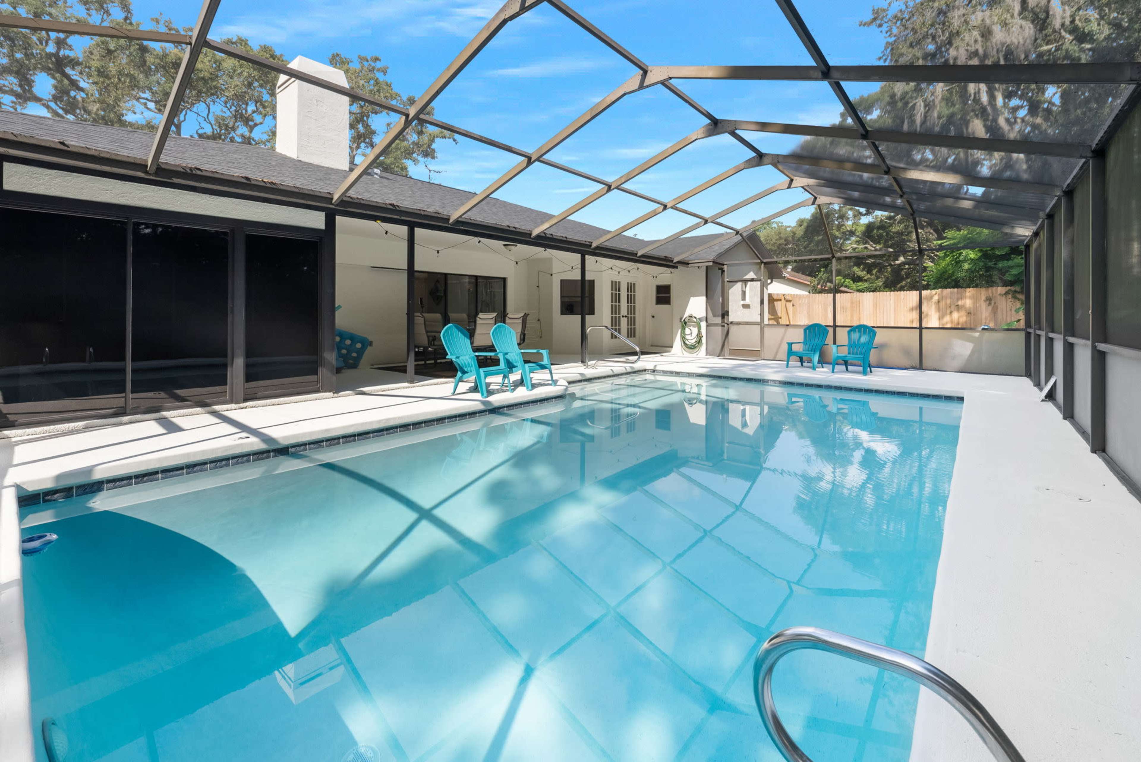 A clear swimming pool is enclosed by a screened patio, with a house and lounge chairs visible in the background.