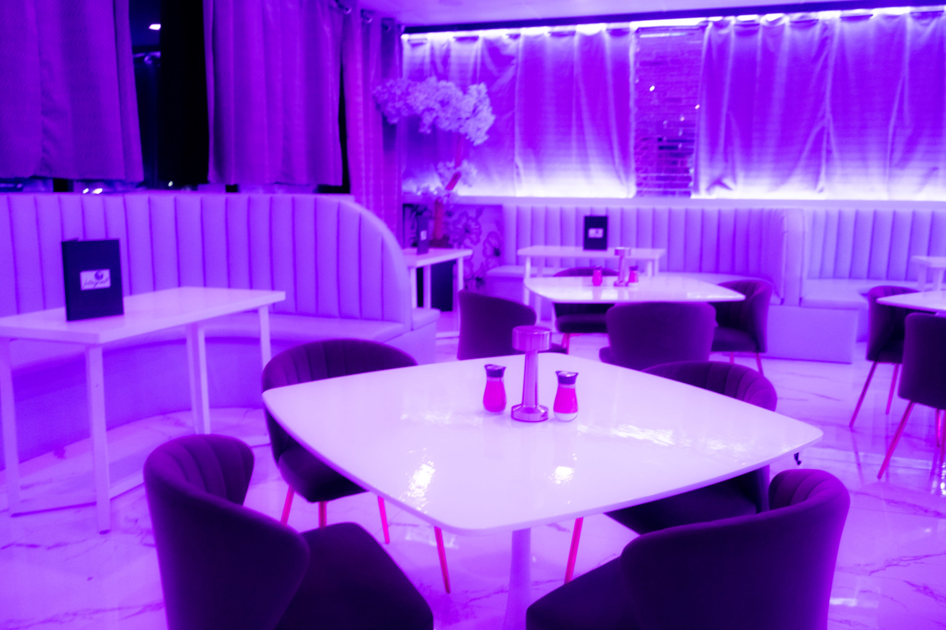 The image shows a modern restaurant interior with white tables, purple upholstered chairs, and ambient purple lighting.