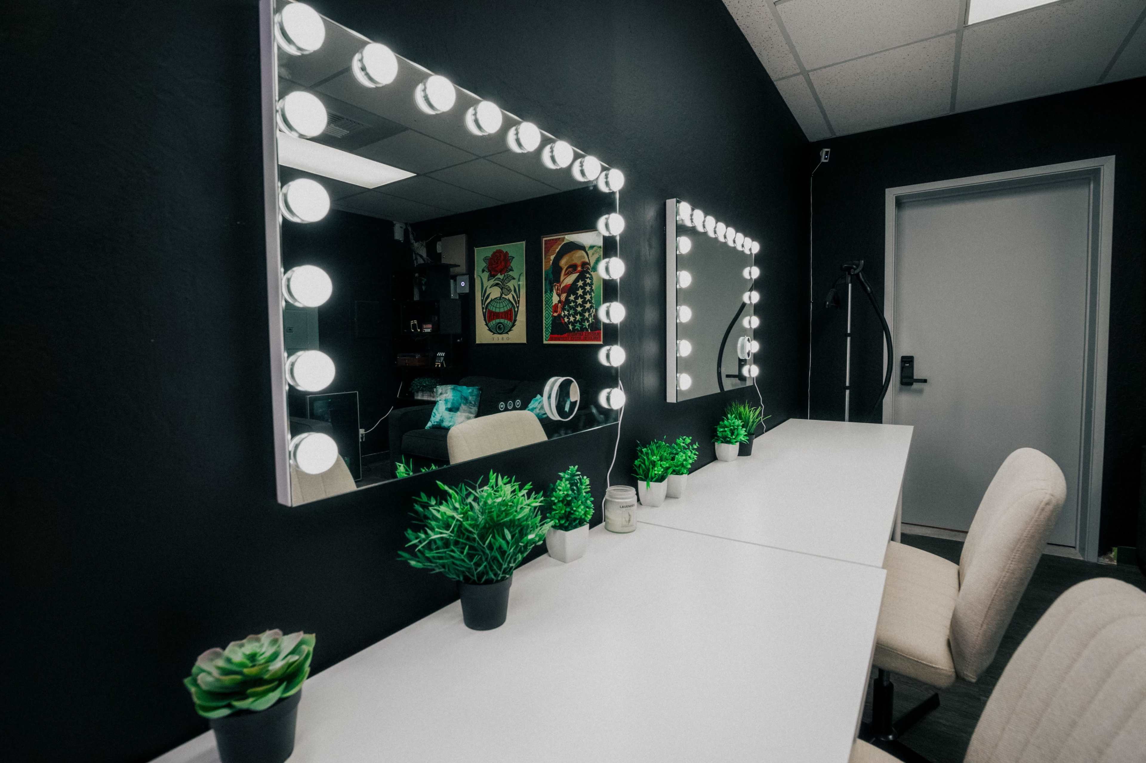 A room with two lighted mirrors above white vanities complemented by small potted plants and a door leading to another area.