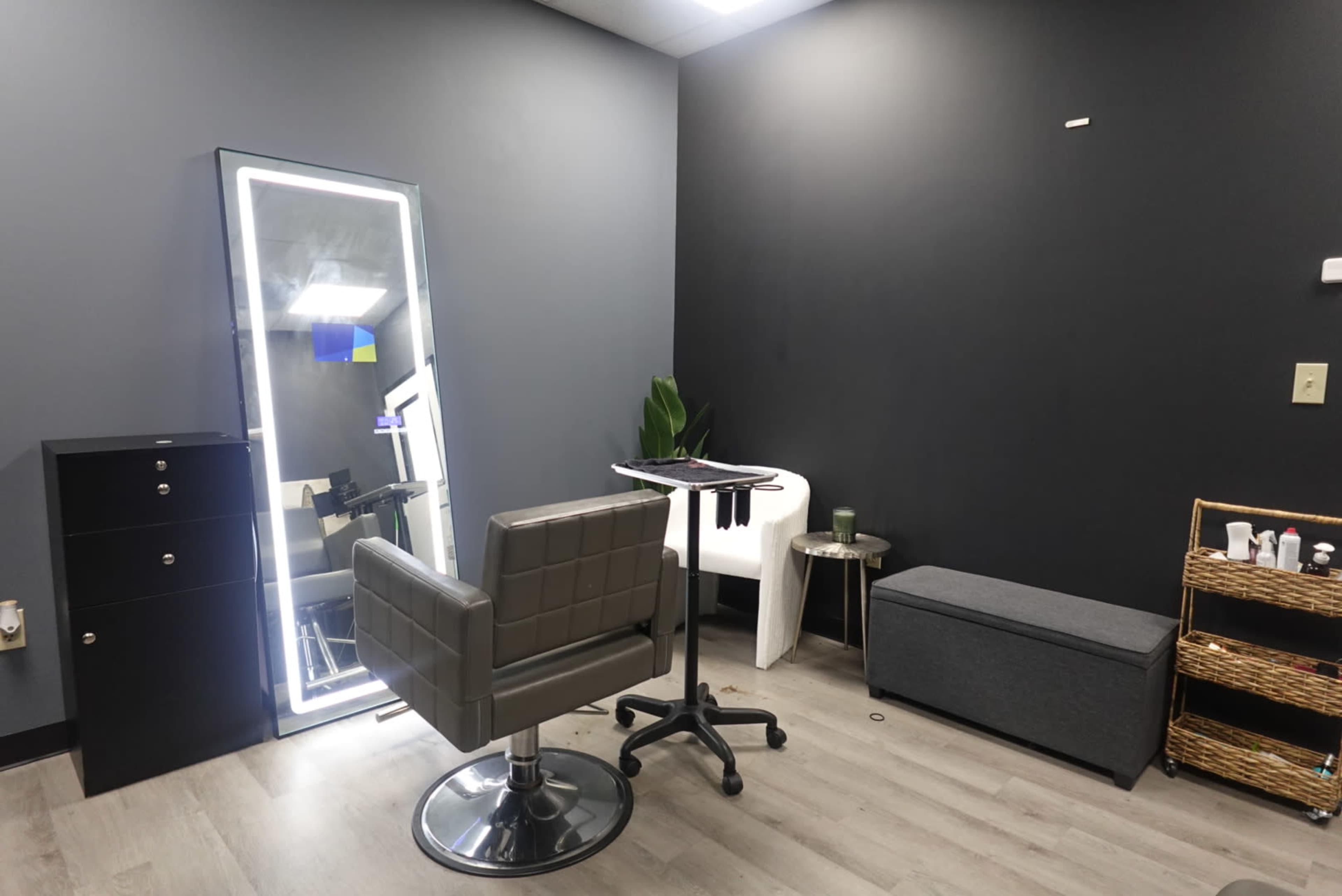 The image shows a modern hair salon interior featuring a stylist chair, a tall mirror, a small table with tools, and storage units against dark walls.