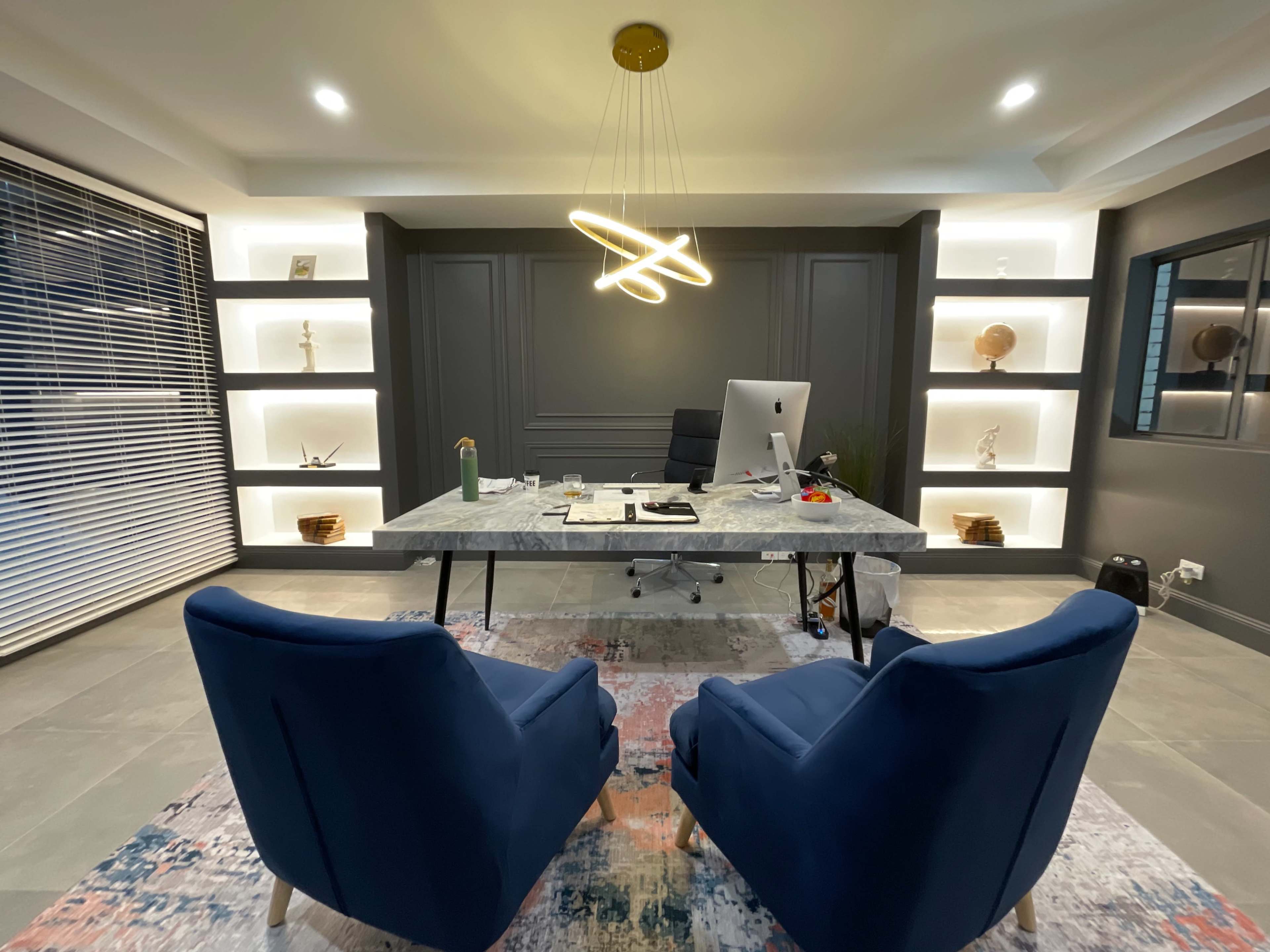 A modern office features a sleek marble desk with an iMac, two blue armchairs, and illuminated shelves displaying decorative items.
