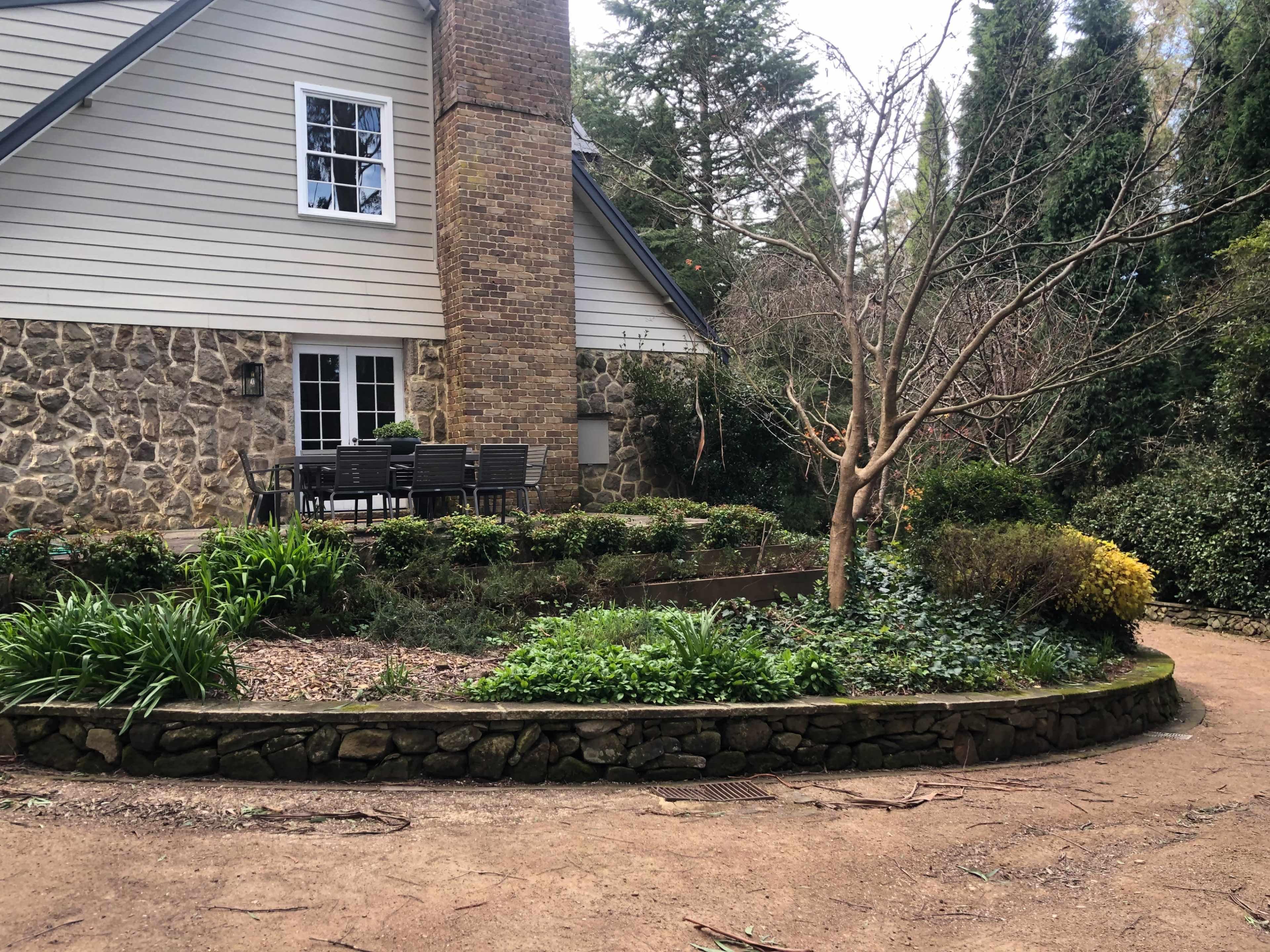 A stone-bordered garden surrounds a small, leafless tree next to a house with a wooden deck and seating area.