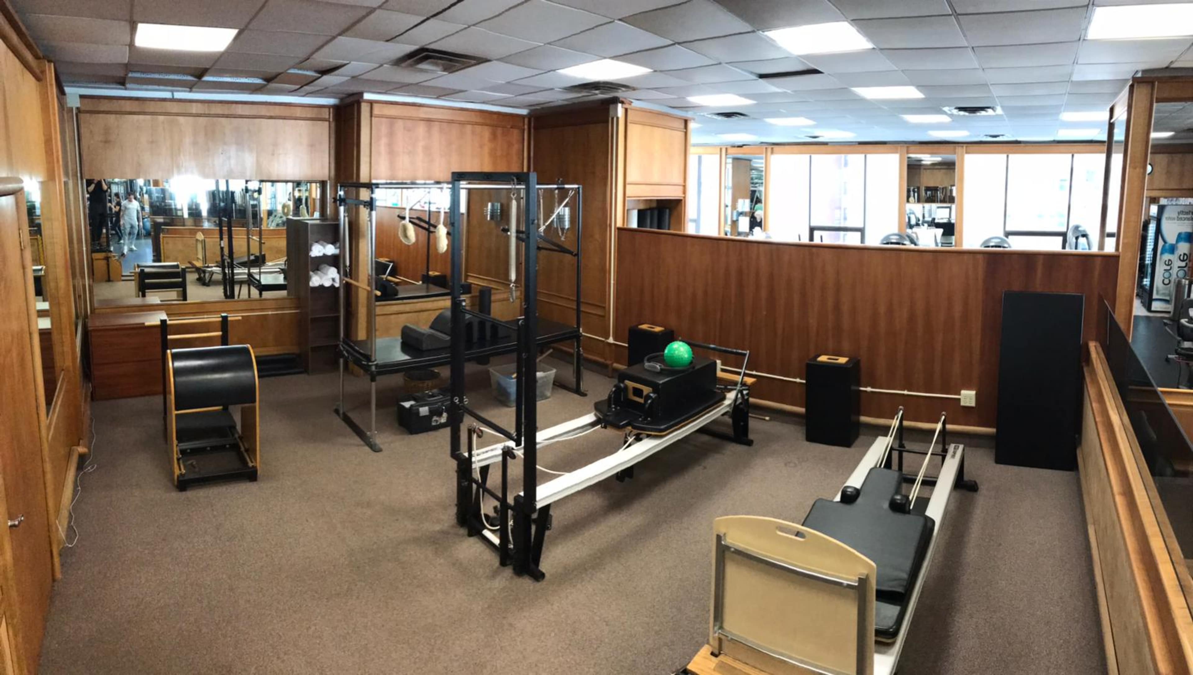 A well-lit Pilates studio equipped with various exercise machines, including a reformer and a cadillac, set against wooden paneling and mirrors.