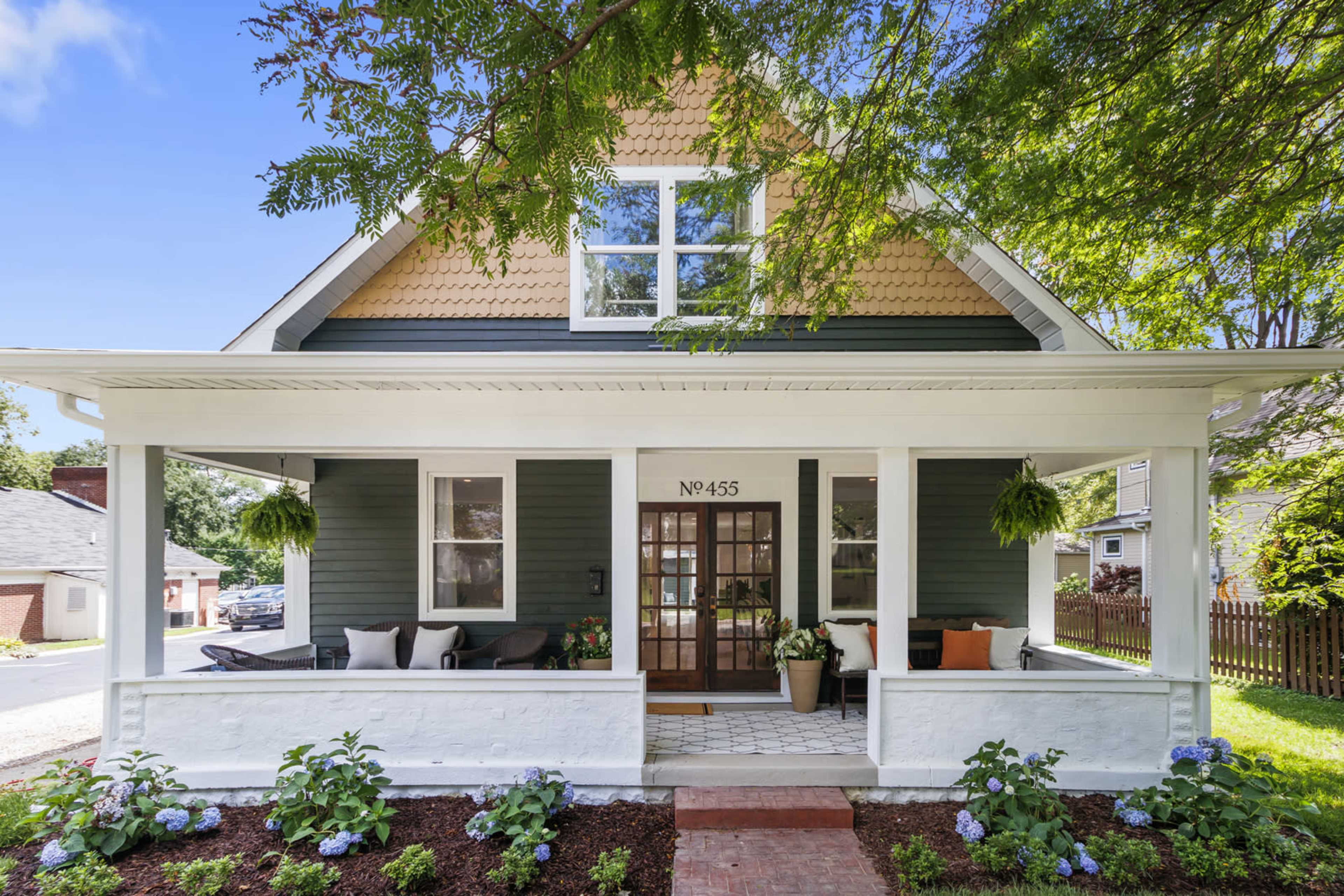 A two-story house with a green exterior, white trim, and a front porch decorated with potted plants and seating.
