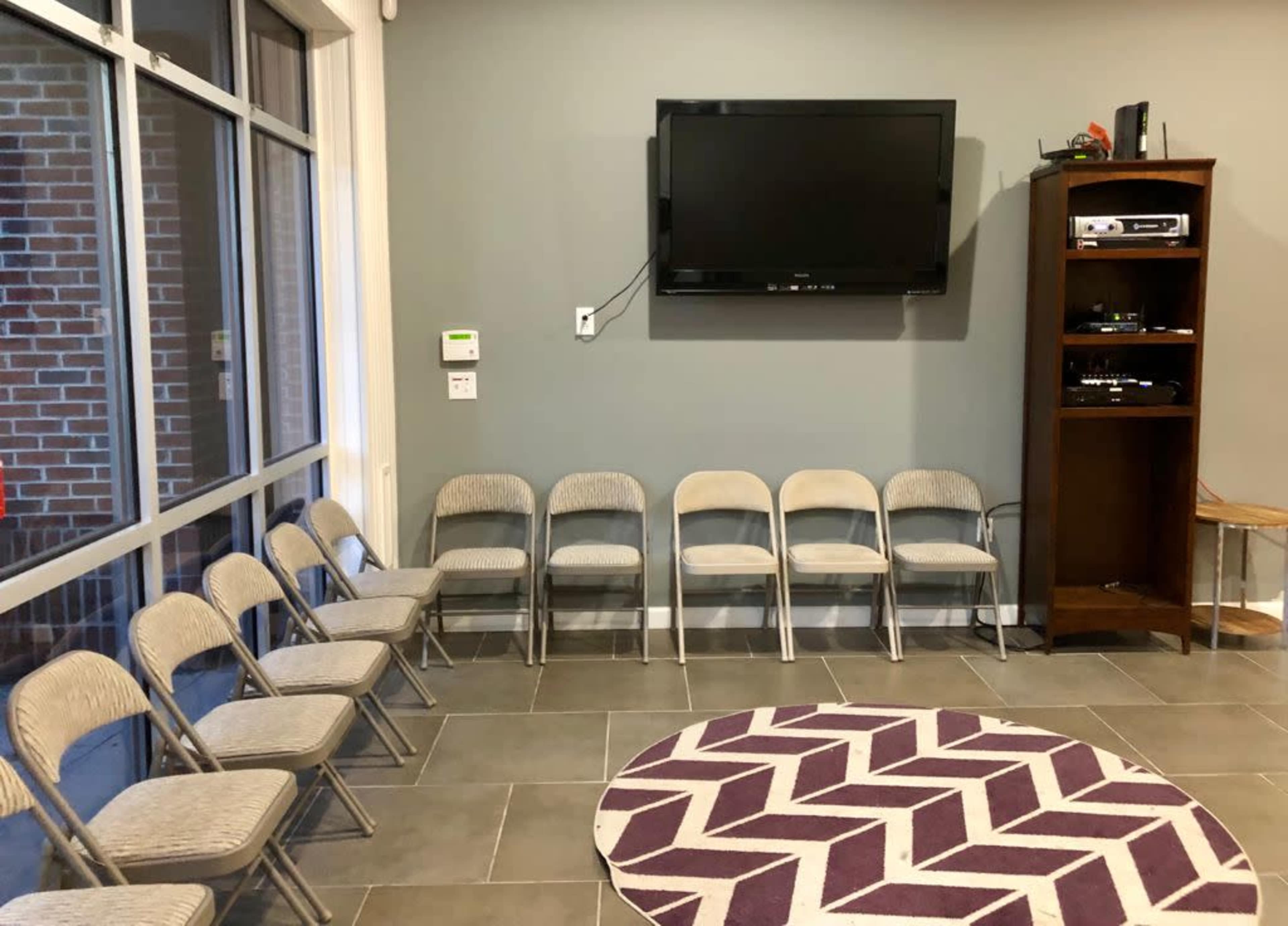 A room with a row of metal folding chairs facing a wall-mounted television, alongside a wooden media stand and a round purple rug on the tiled floor.