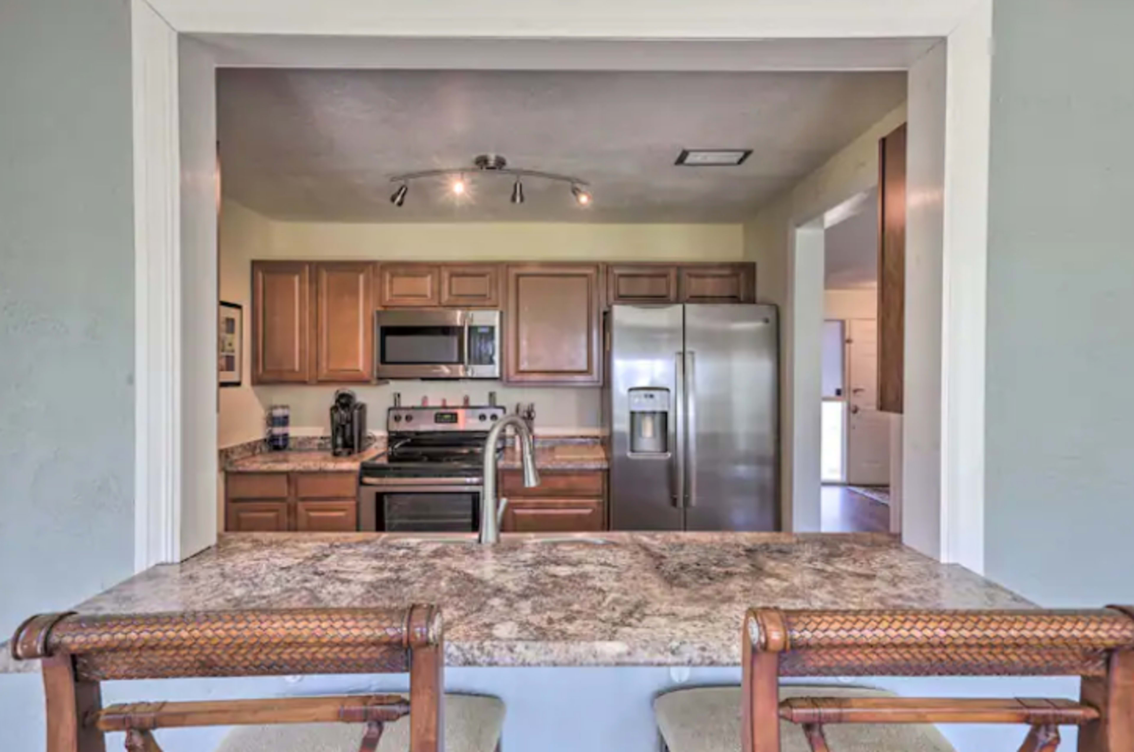 A kitchen viewed from a bar area, featuring wooden cabinets, stainless steel appliances, and a granite countertop.