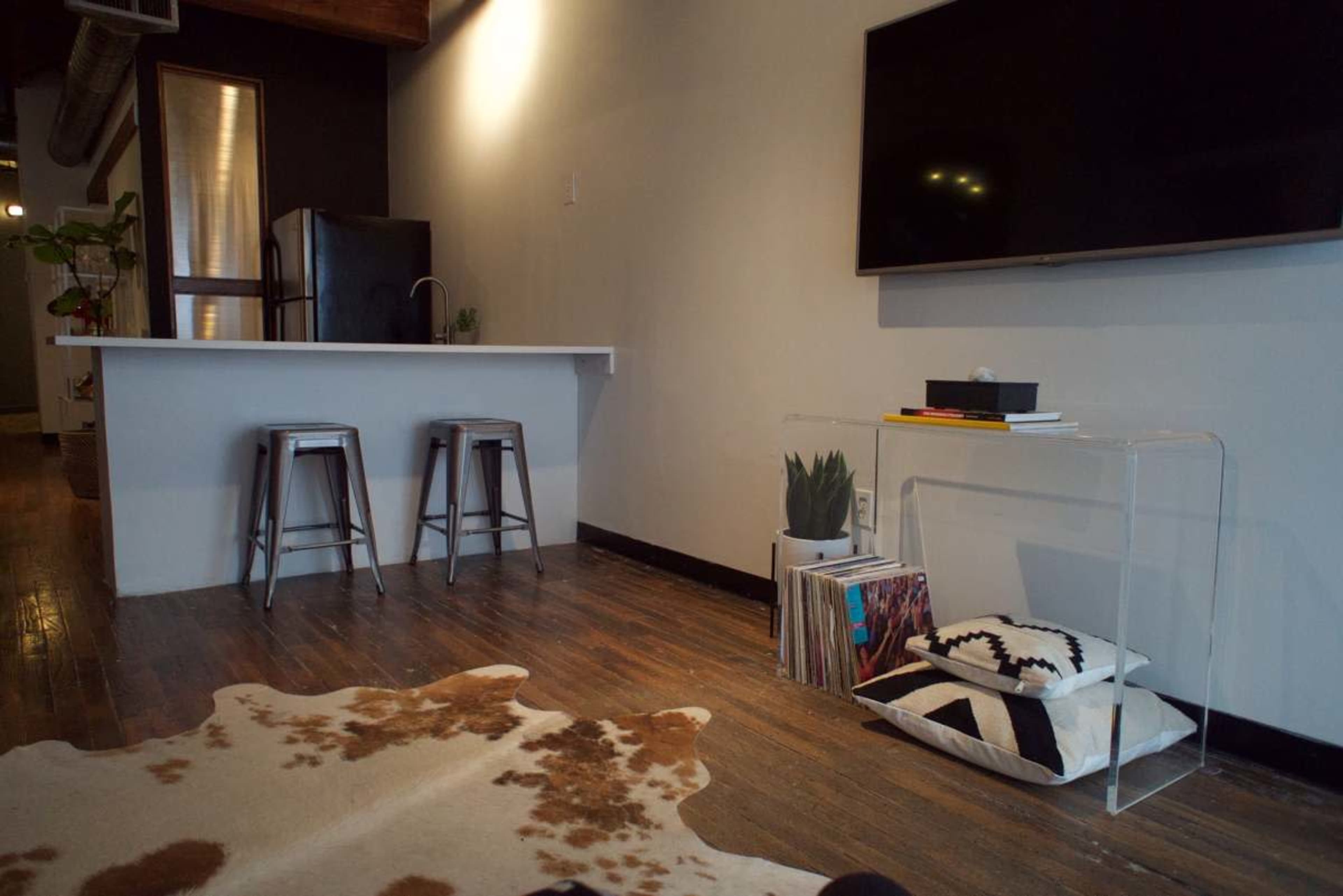 The image shows a modern living space with a small kitchen area featuring two stools, a television mounted on the wall, and a clear acrylic table displaying books and decorative items, all set on hardwood floors.