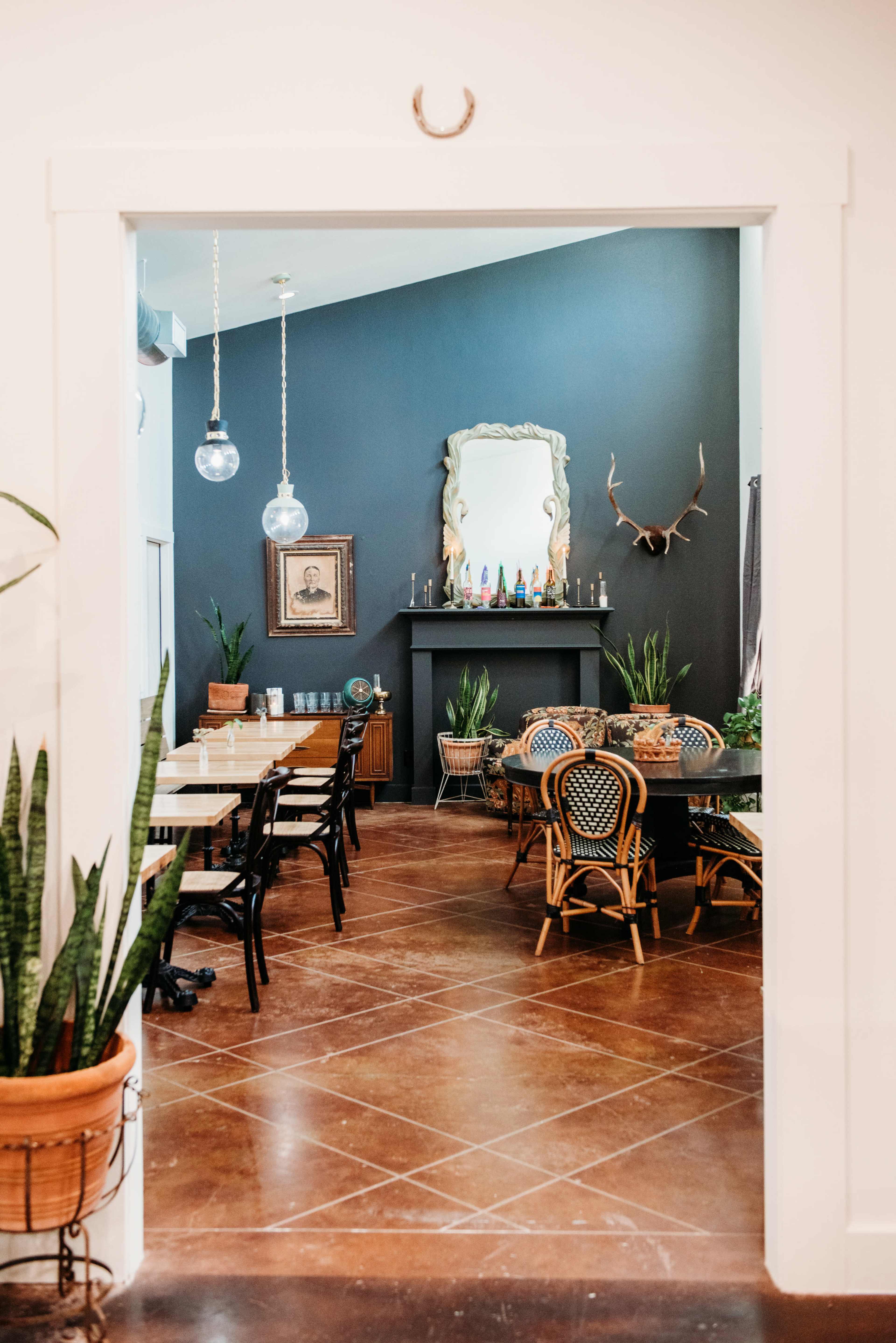 A dining area with wooden tables and chairs, decorative plants, and a dark wall featuring a mirror and antlers.