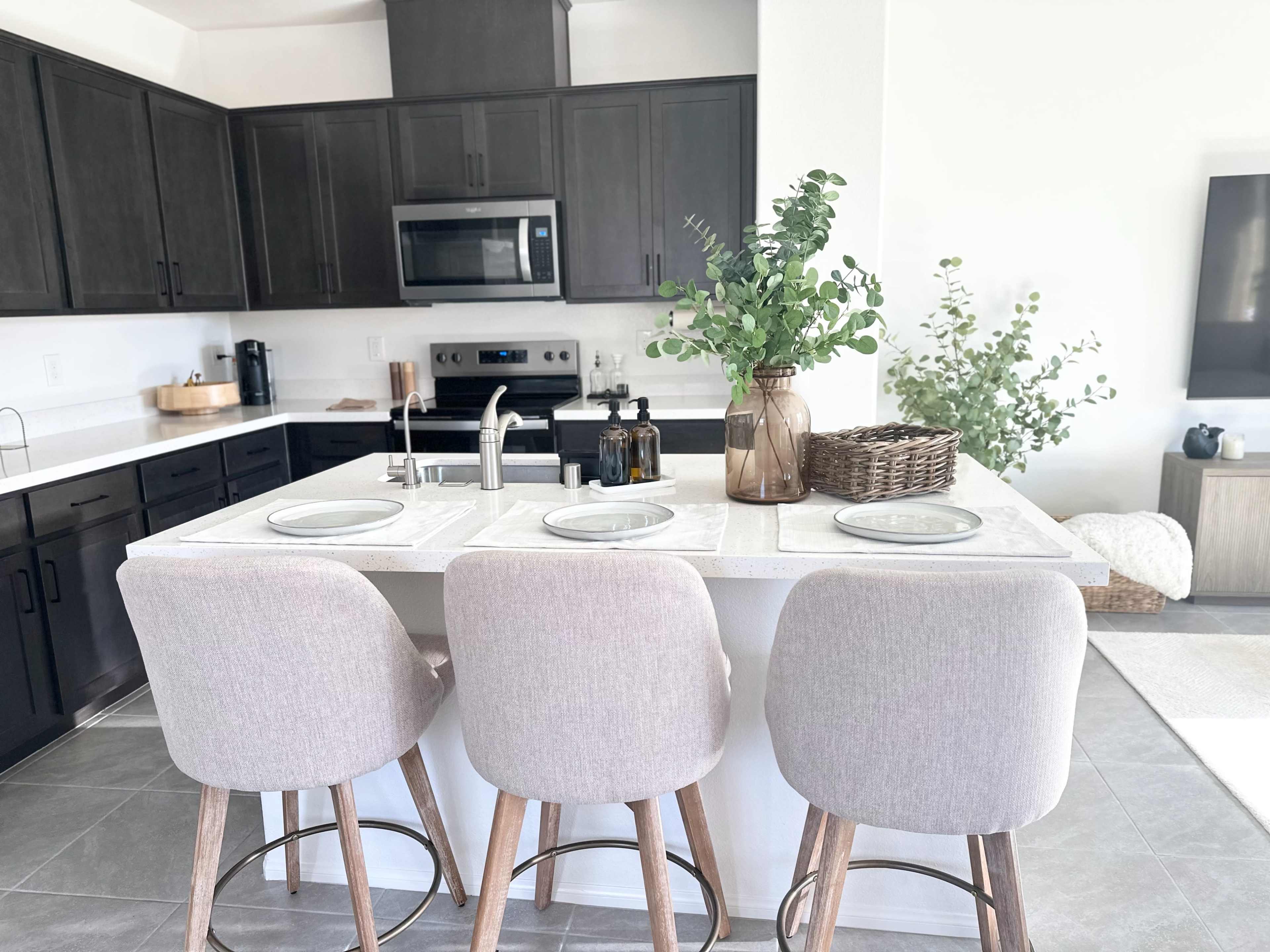 A modern kitchen with dark cabinetry, a central island with three beige bar stools, and decorative plants on the counter.