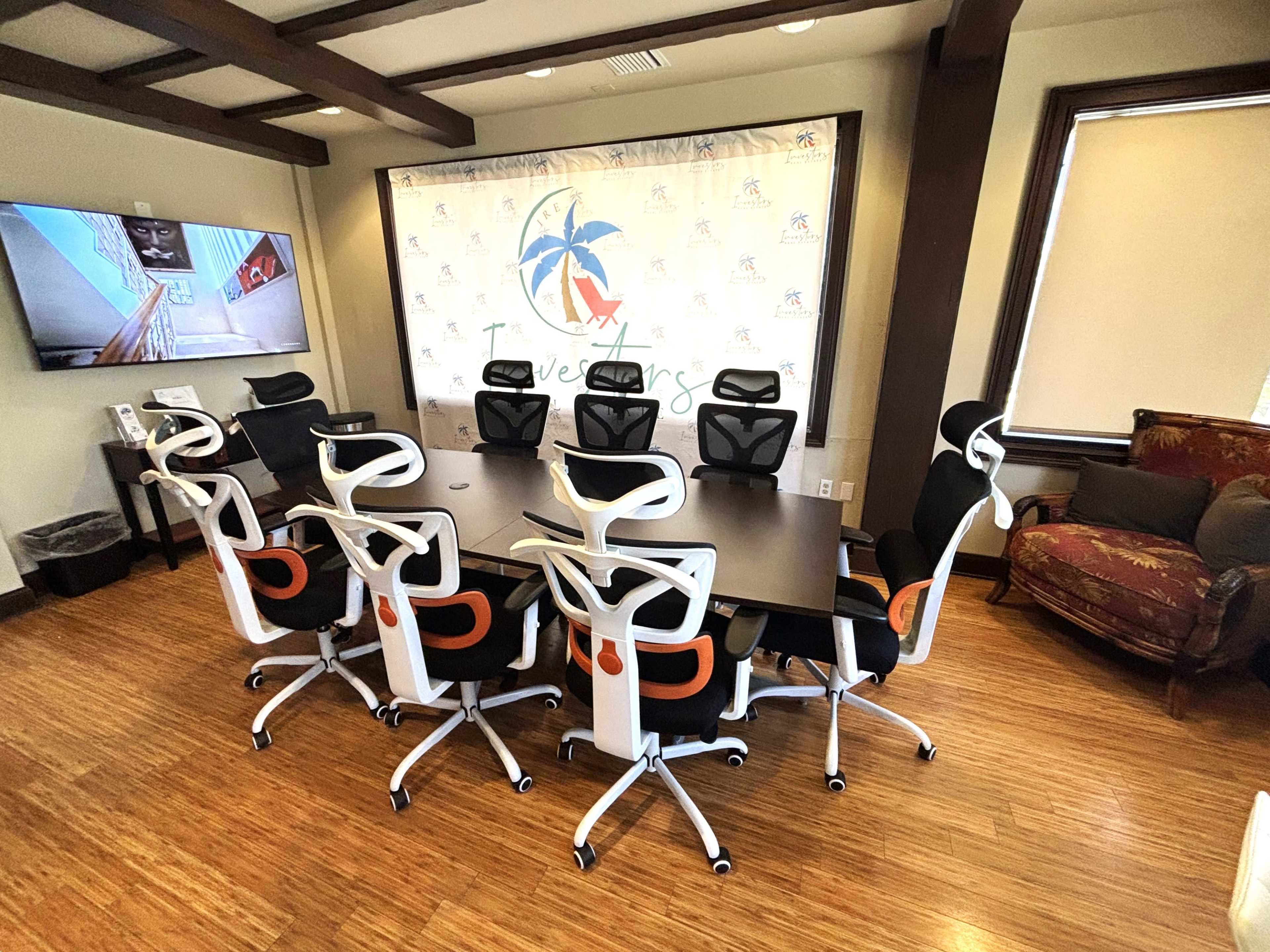 A modern conference room features a long table surrounded by ergonomic chairs and a large screen displaying a logo.