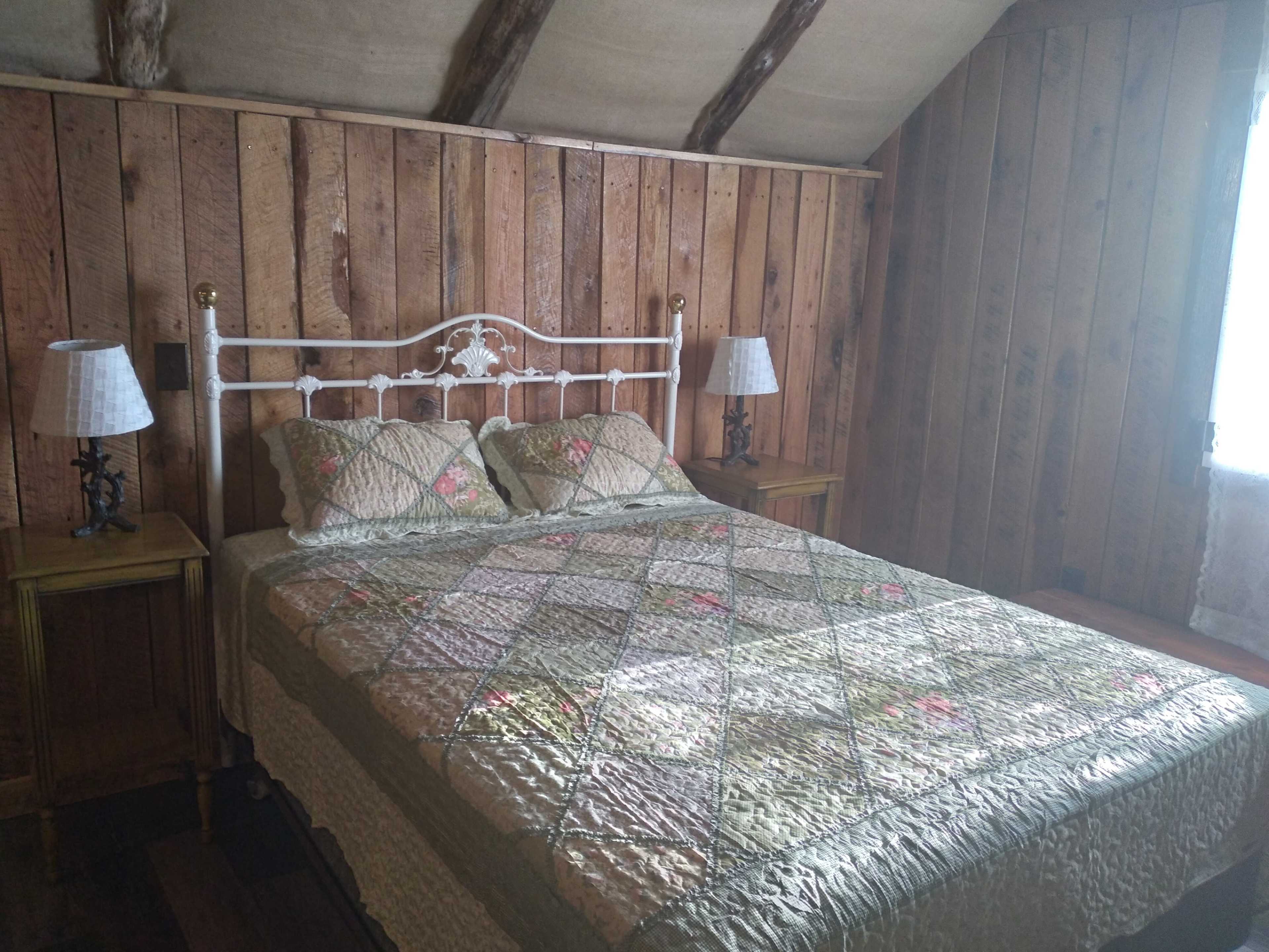 The image shows a wooden bedroom with a bed covered in a patterned quilt and two matching bedside lamps on small tables.