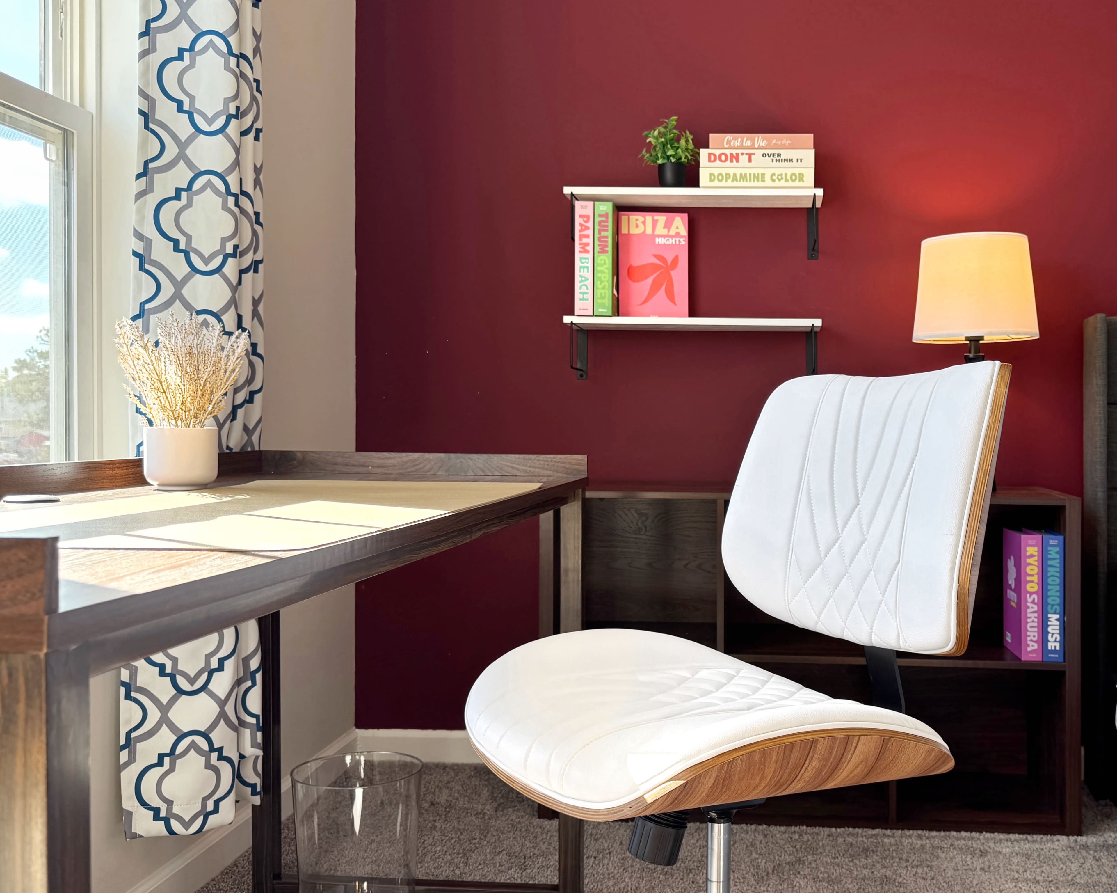 The image shows a modern home office setup featuring a white chair at a desk, with a wall shelf displaying colorful books and a lamp on a red wall.