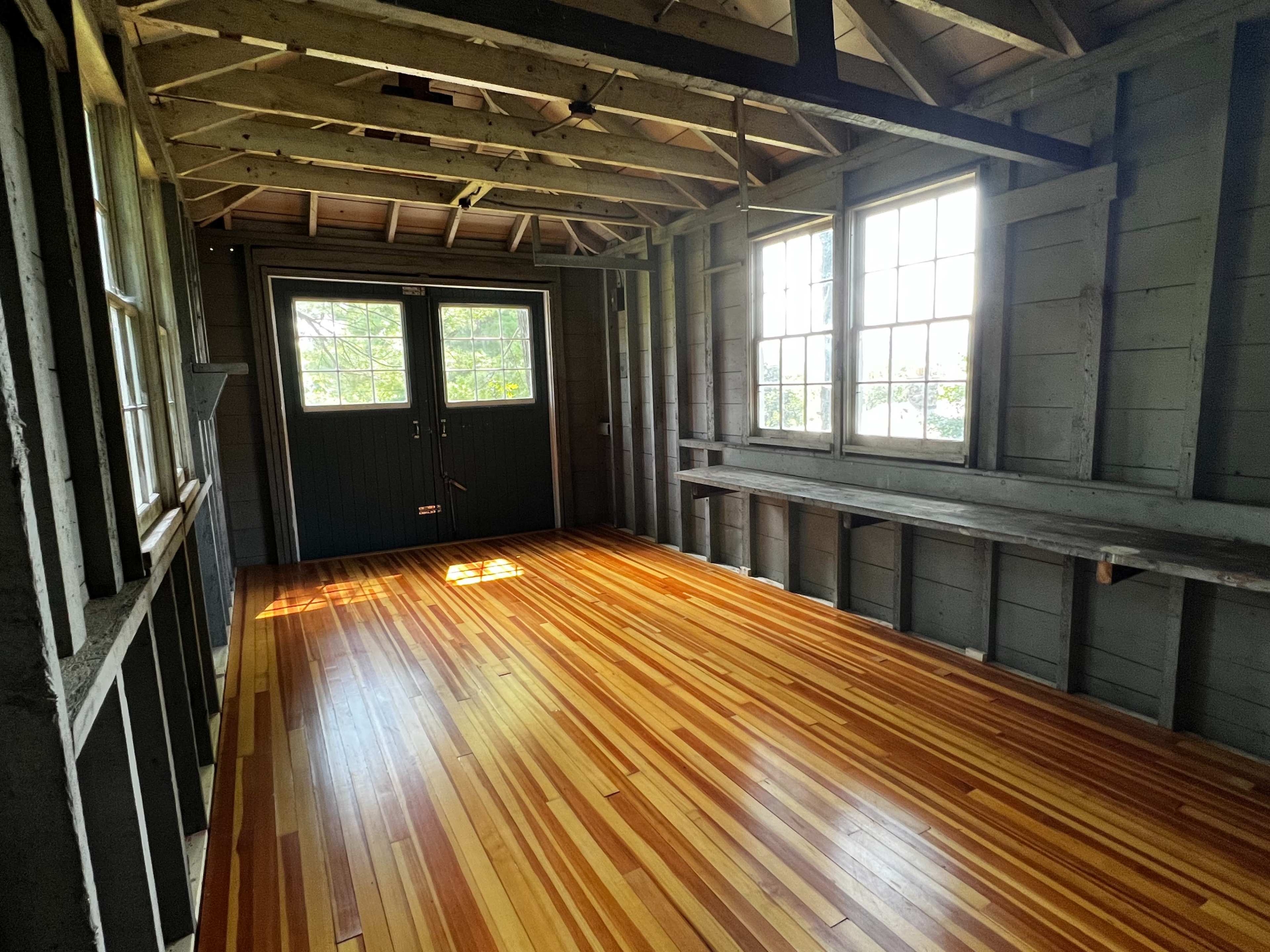 A partially finished room with wooden floors, exposed wall framing, and two windows letting in natural light.